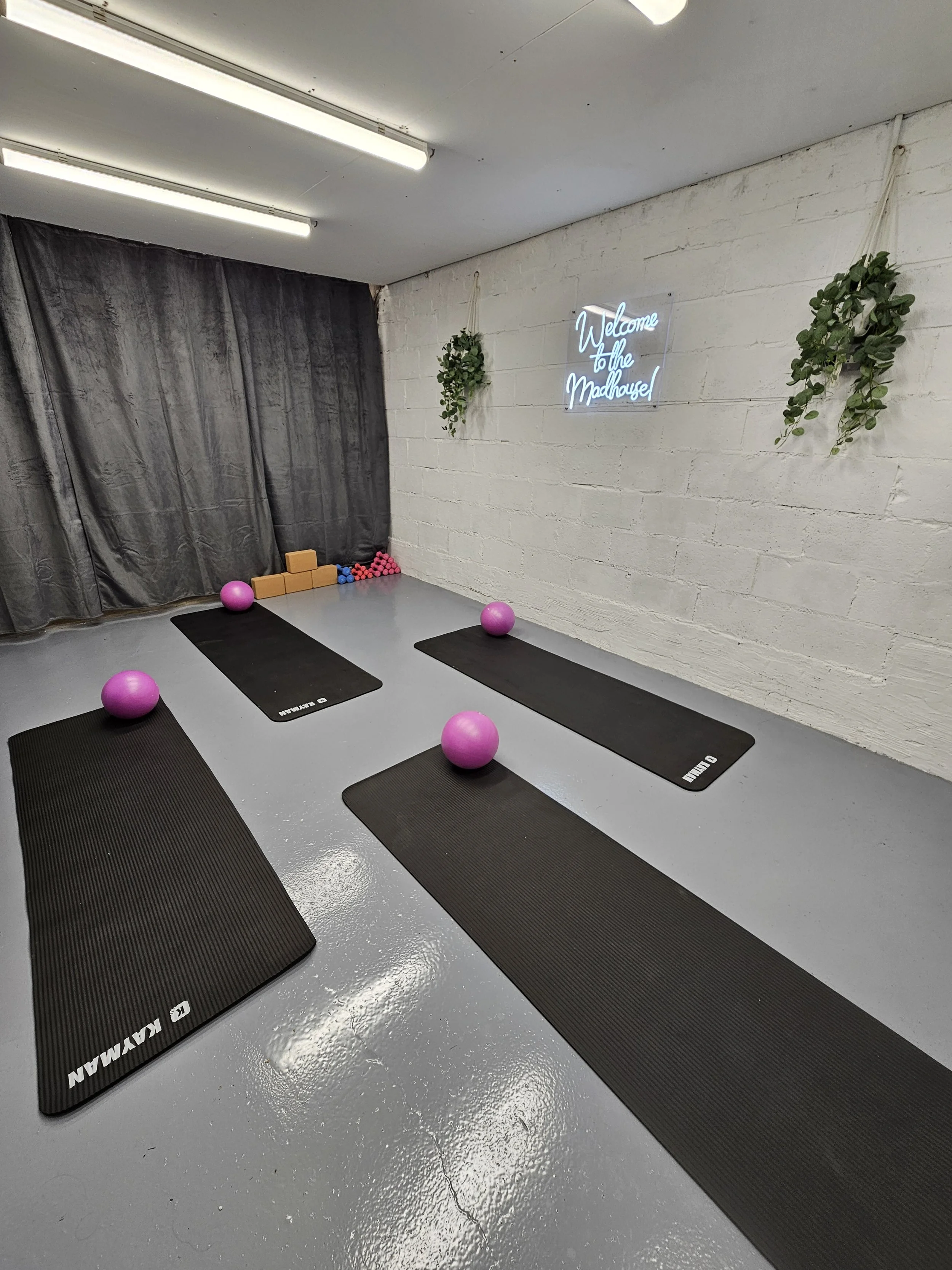Indoor yoga studio with three black yoga mats, purple exercise balls, and pink hand weights. Two hanging plants, a neon sign that reads 'Welcome to the Madhouse', and a black curtain in the background.