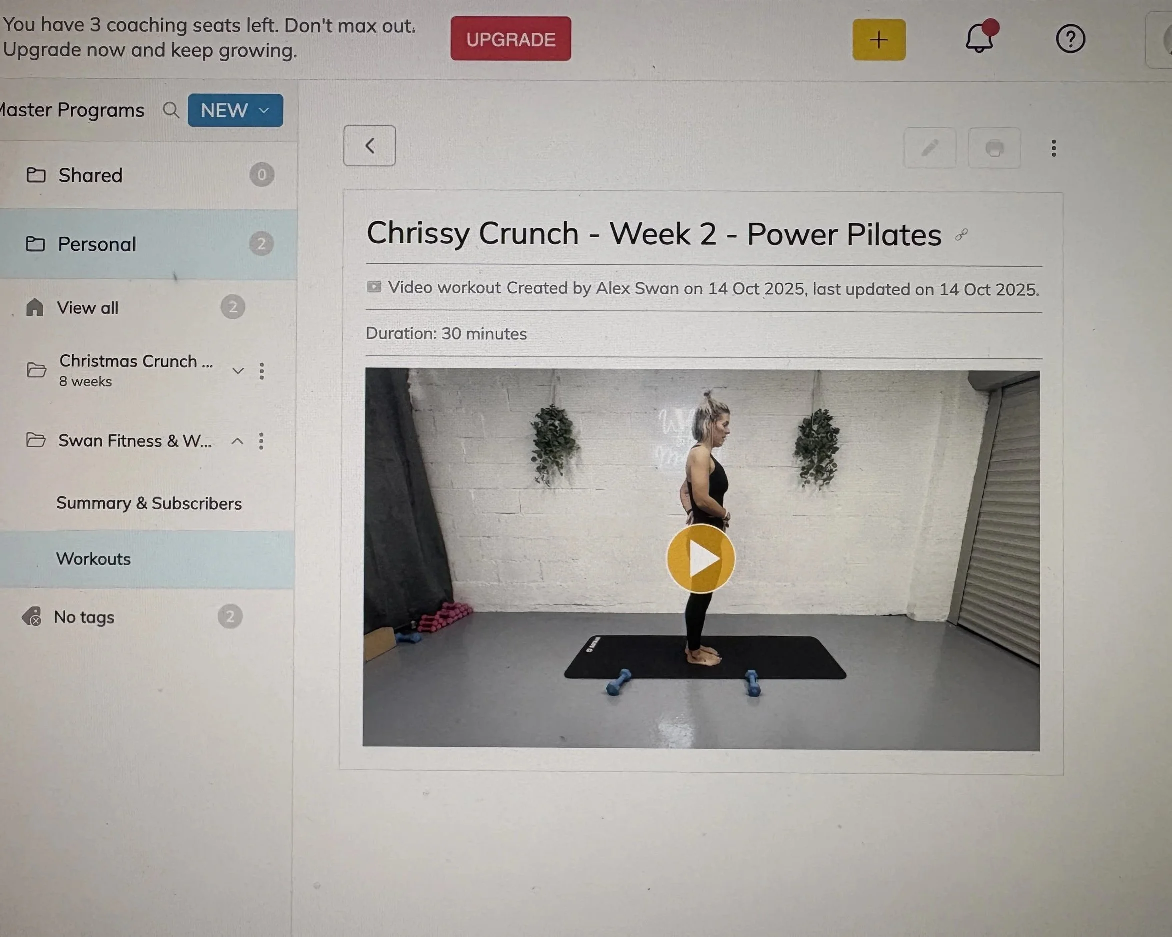 Screenshot of a workout video titled 'Chrissy Crunch - Week 2 - Power Pilates' showing a woman standing on a yoga mat with weights on either side in a studio with white brick walls and hanging plants.