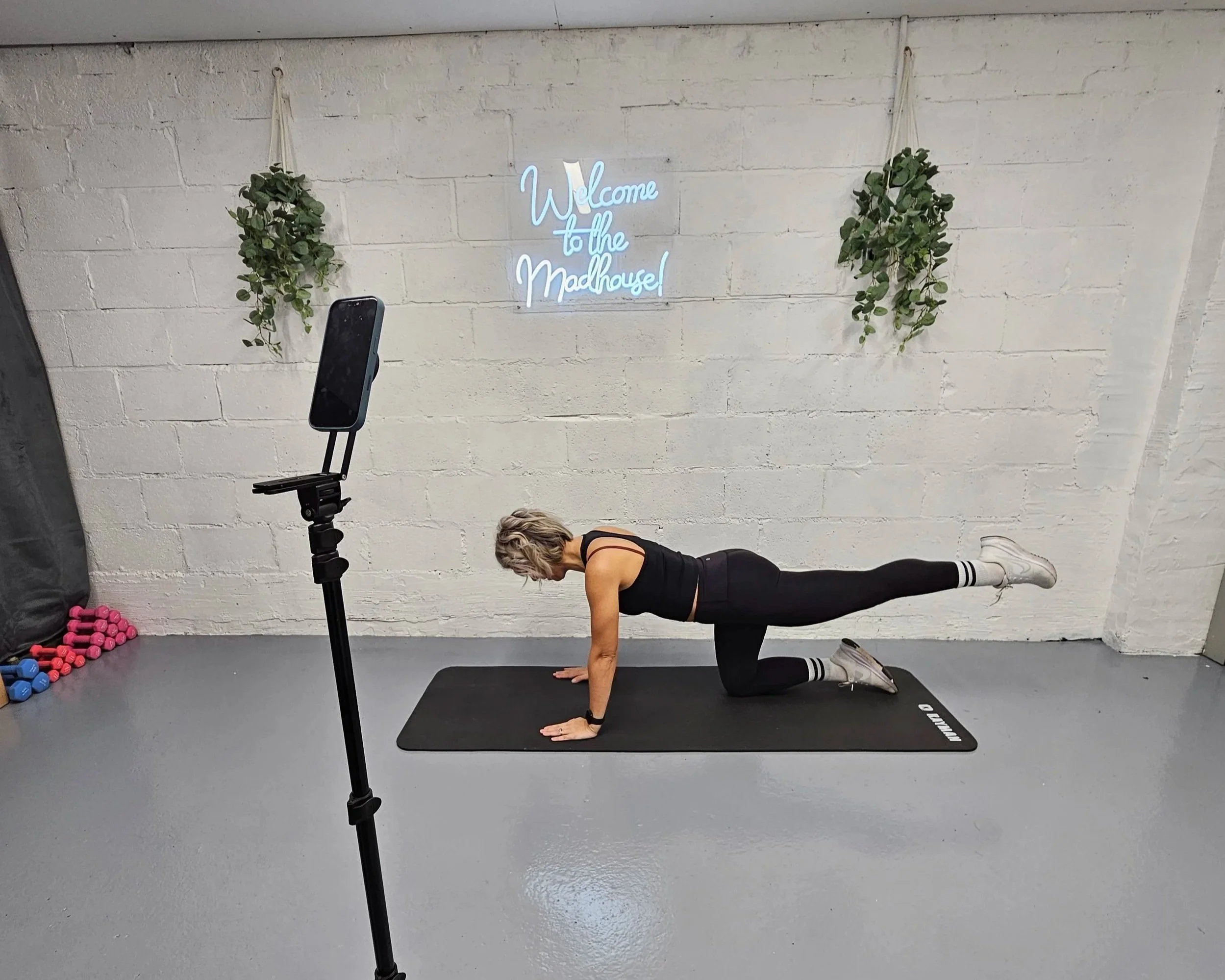 A woman is doing a plank exercise on a black mat in a gym, with a smartphone on a tripod in front of her capturing the workout. She is wearing a black tank top, black leggings, and white shoes. The background features a white brick wall with a neon sign that reads "Welcome to the Madhouse!" and hanging green plants on either side.