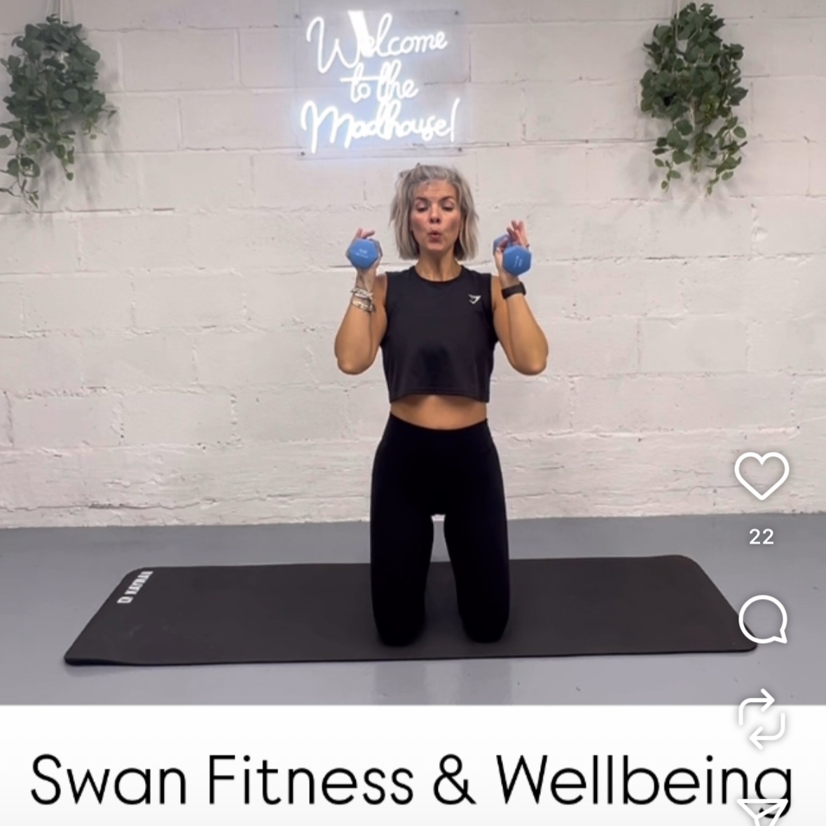 A woman kneeling on a yoga mat in a fitness studio, holding blue dumbbells in both hands at shoulder height, with a neon sign on the wall behind her that says "Welcome to the Mad House!" and green plants on either side.