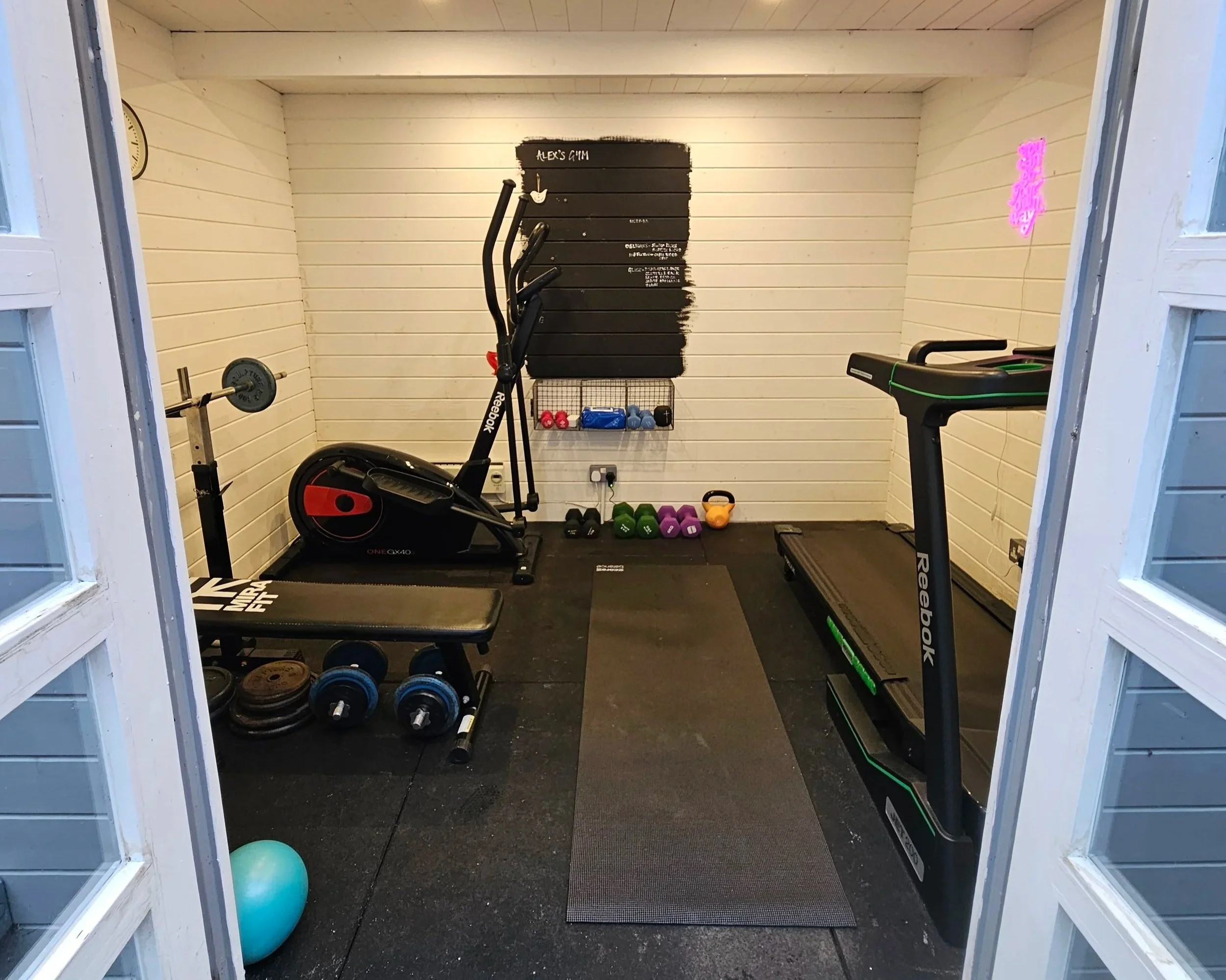 Home gym with cardio equipment, free weights, dumbbells, exercise ball, and workout accessories inside a small room with white panel walls.