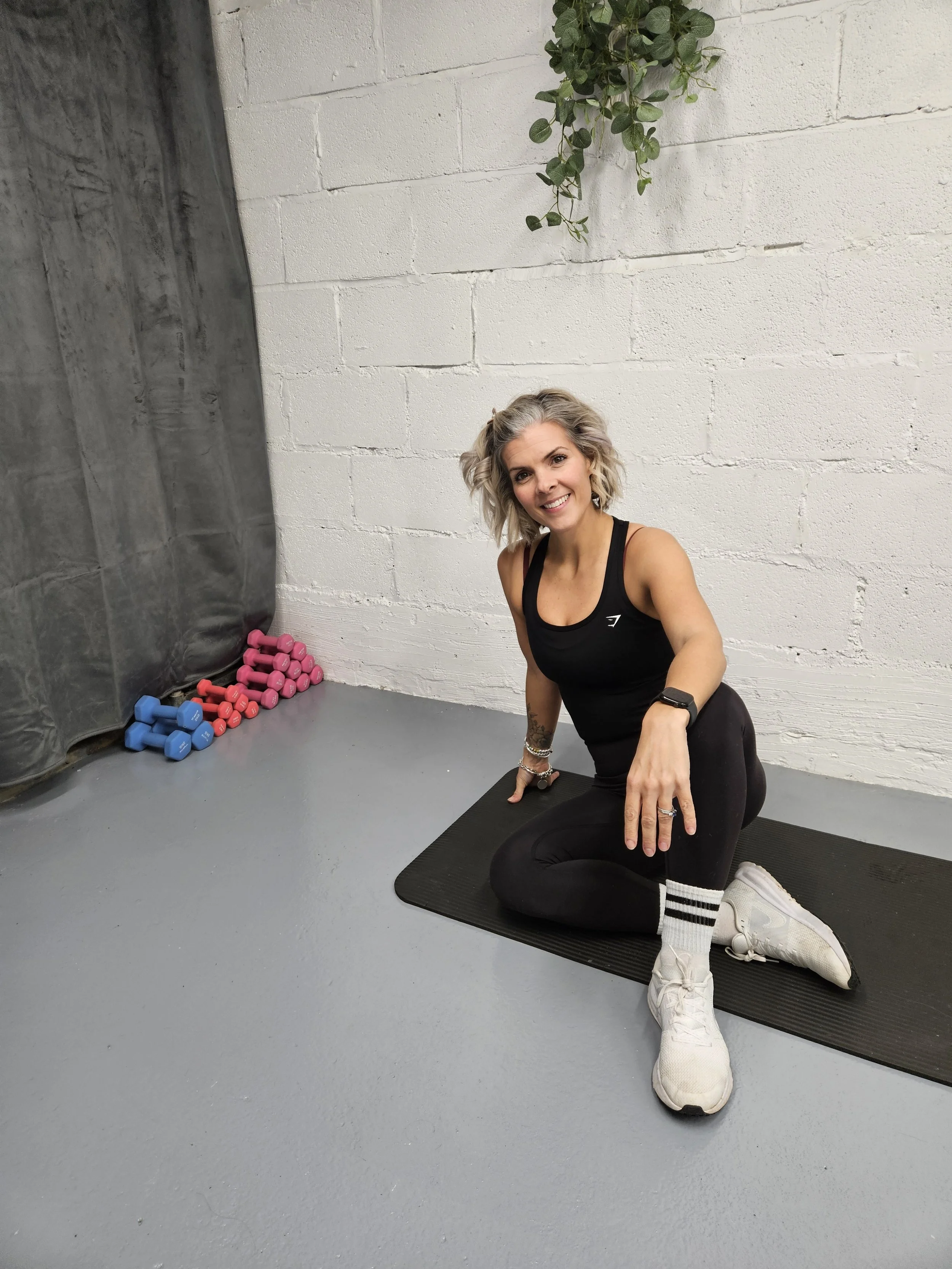 A woman with blonde wavy hair kneeling on a black exercise mat in a gym, smiling at the camera. She is wearing a black tank top, black leggings, white sneakers, and striped socks. There are pink and blue dumbbells stacked in the corner and a white brick wall with a hanging green plant in the background.