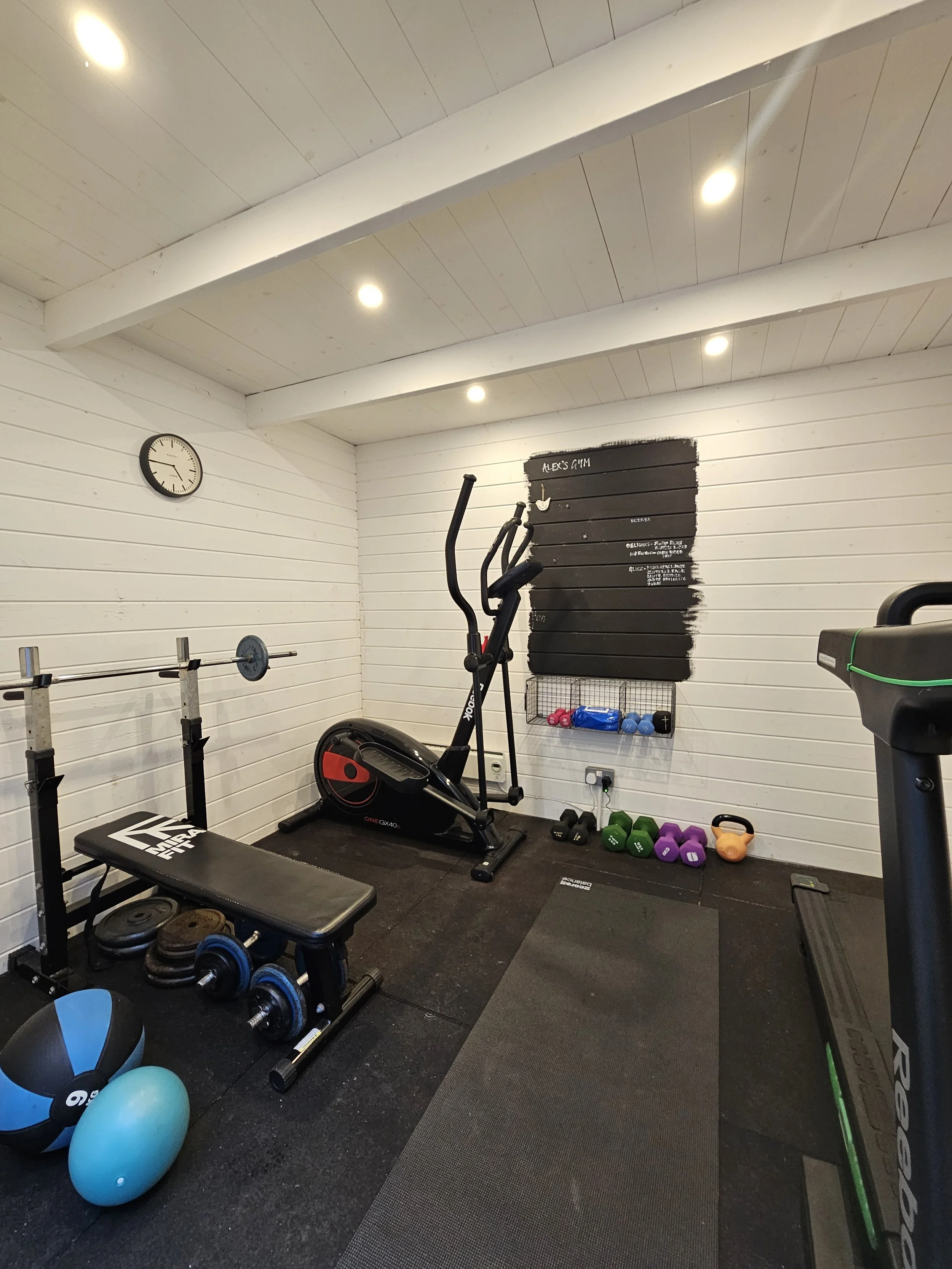 Home gym with white wooden paneled walls, a clock on the wall, black rubber flooring, a bench, dumbbells, medicine balls, a rowing machine, a small shelf with kettlebells, and a black painted wooden board with white lettering.