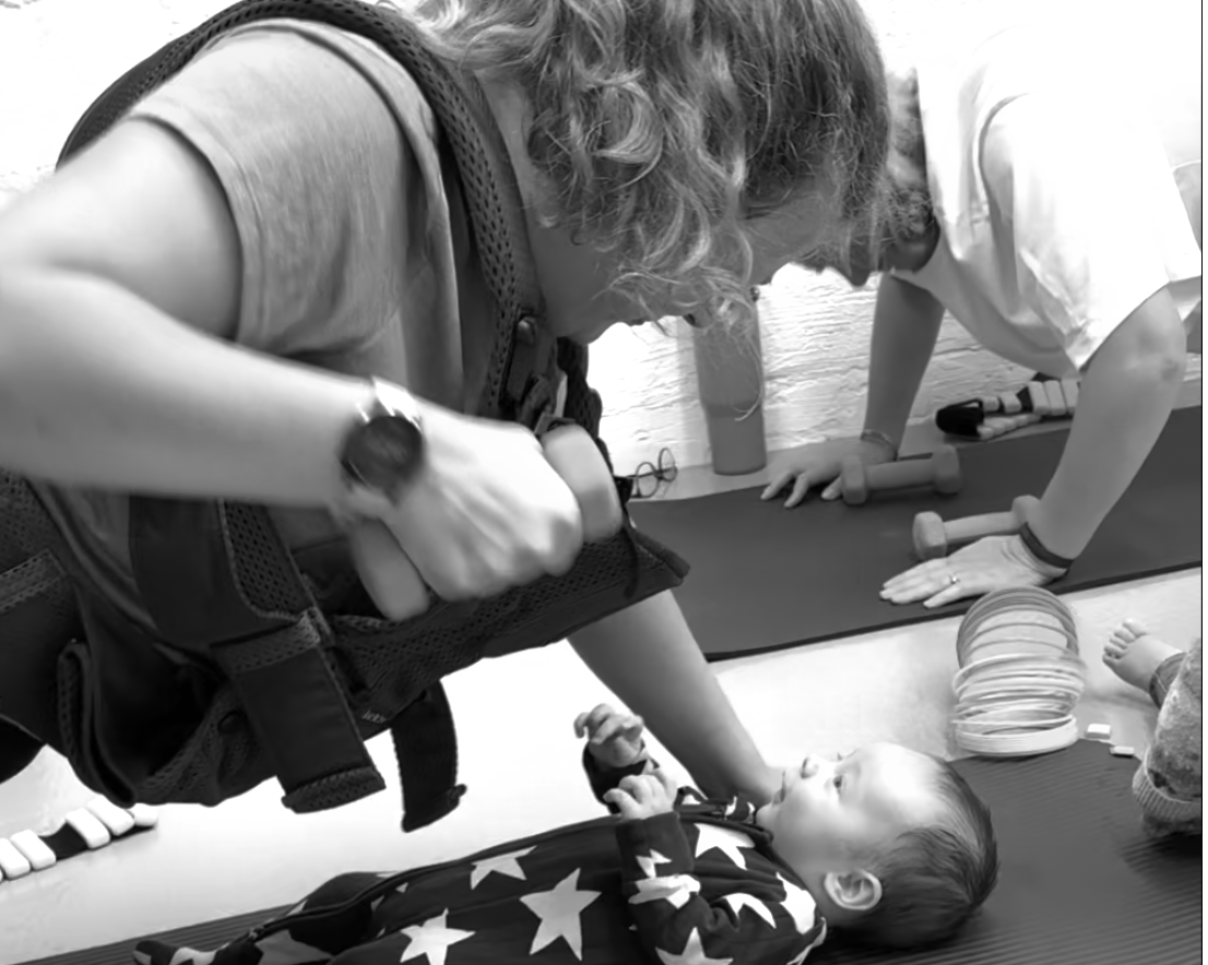 A woman performs a rescue training drill on a baby mannequin, while another person assists by holding a tool, both on a yoga mat.
