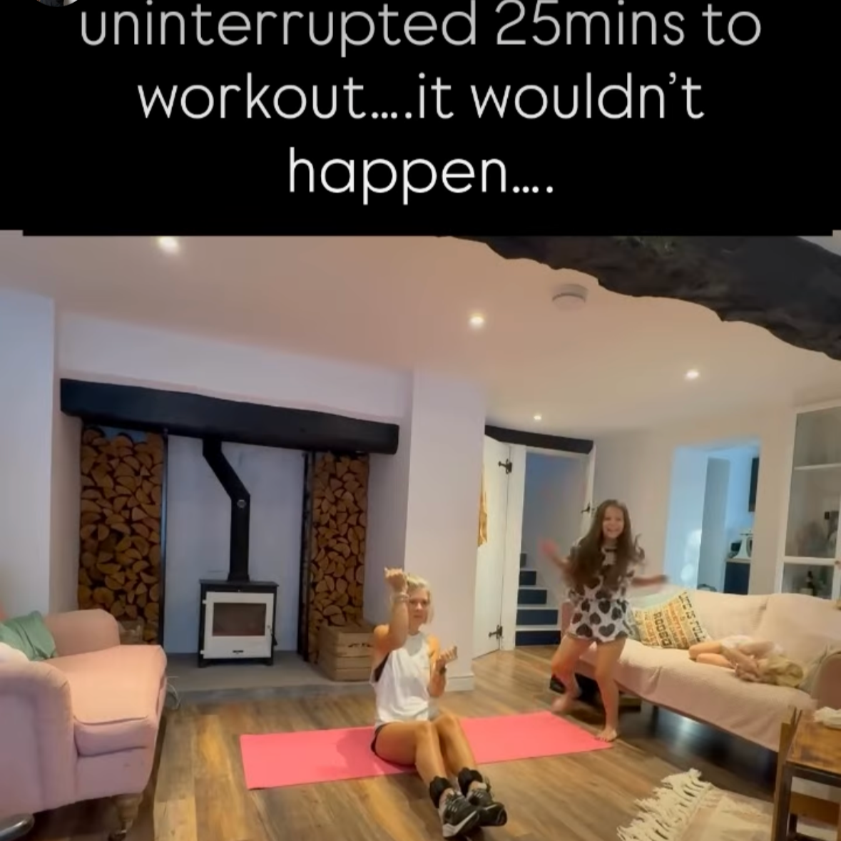 Two young girls are in a living room engaging in a workout or dance activity, with one girl sitting on a pink exercise mat and the other standing and dancing. The room has a wood stove, stacked firewood, a beige sofa with colorful pillows, a pink armchair, and a partially visible dog sleeping on the sofa. The ceiling is white with recessed lights, and there is a doorway in the background.