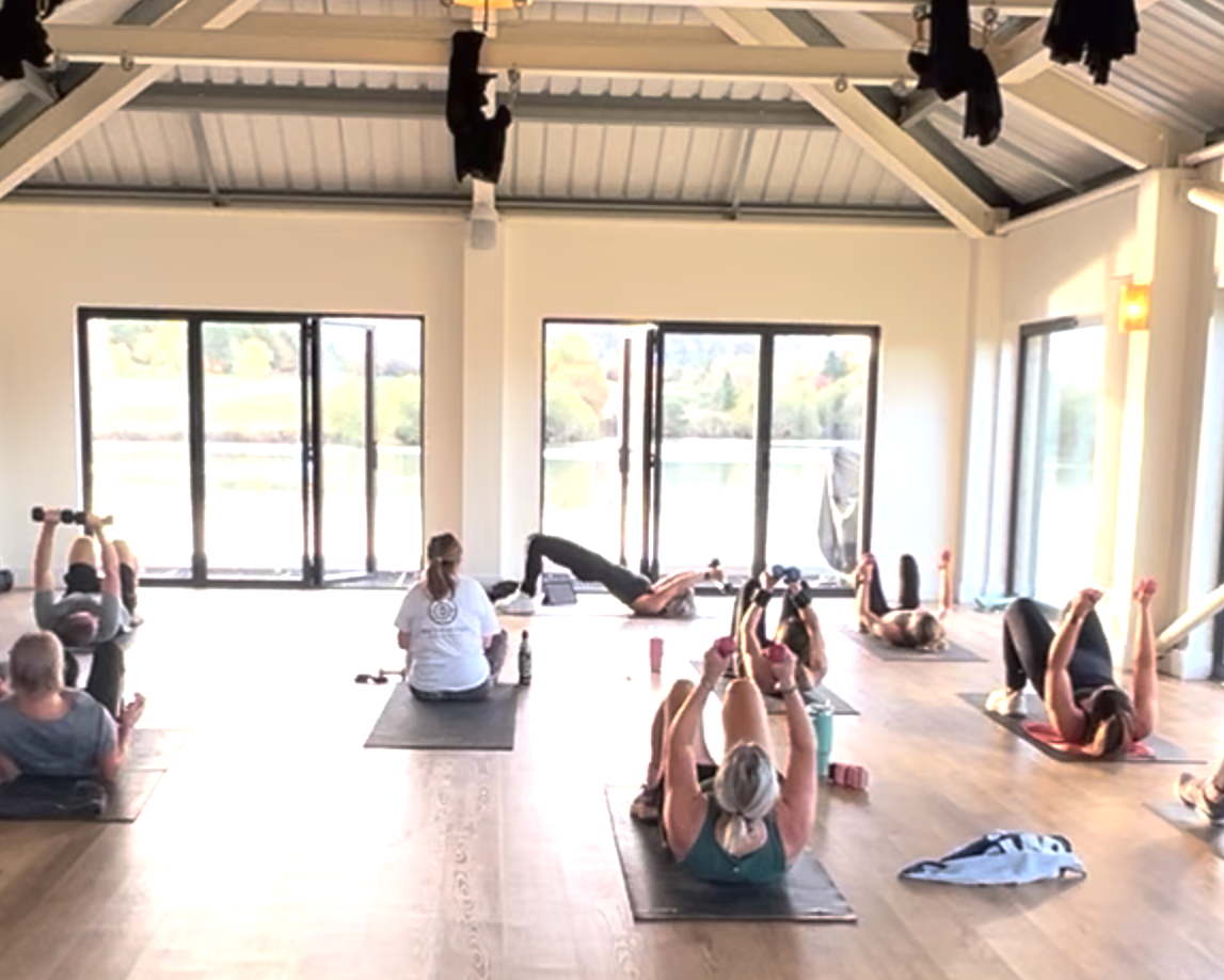 People participating in a yoga class in a bright, airy studio with large windows showing a lakeside view.