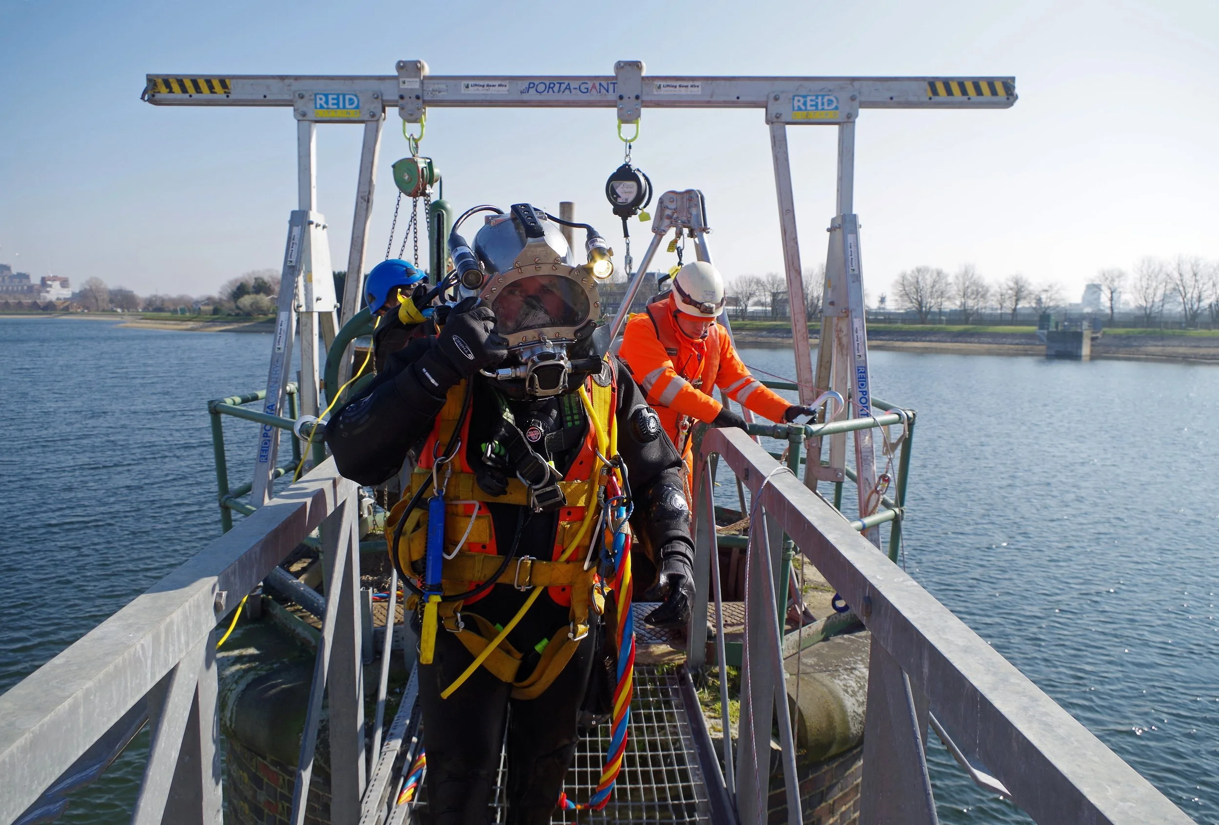 Three workers in safety gear conducting underwater maintenance or inspection on a water intake pipe at a waterway, with a body of water and trees in the background.