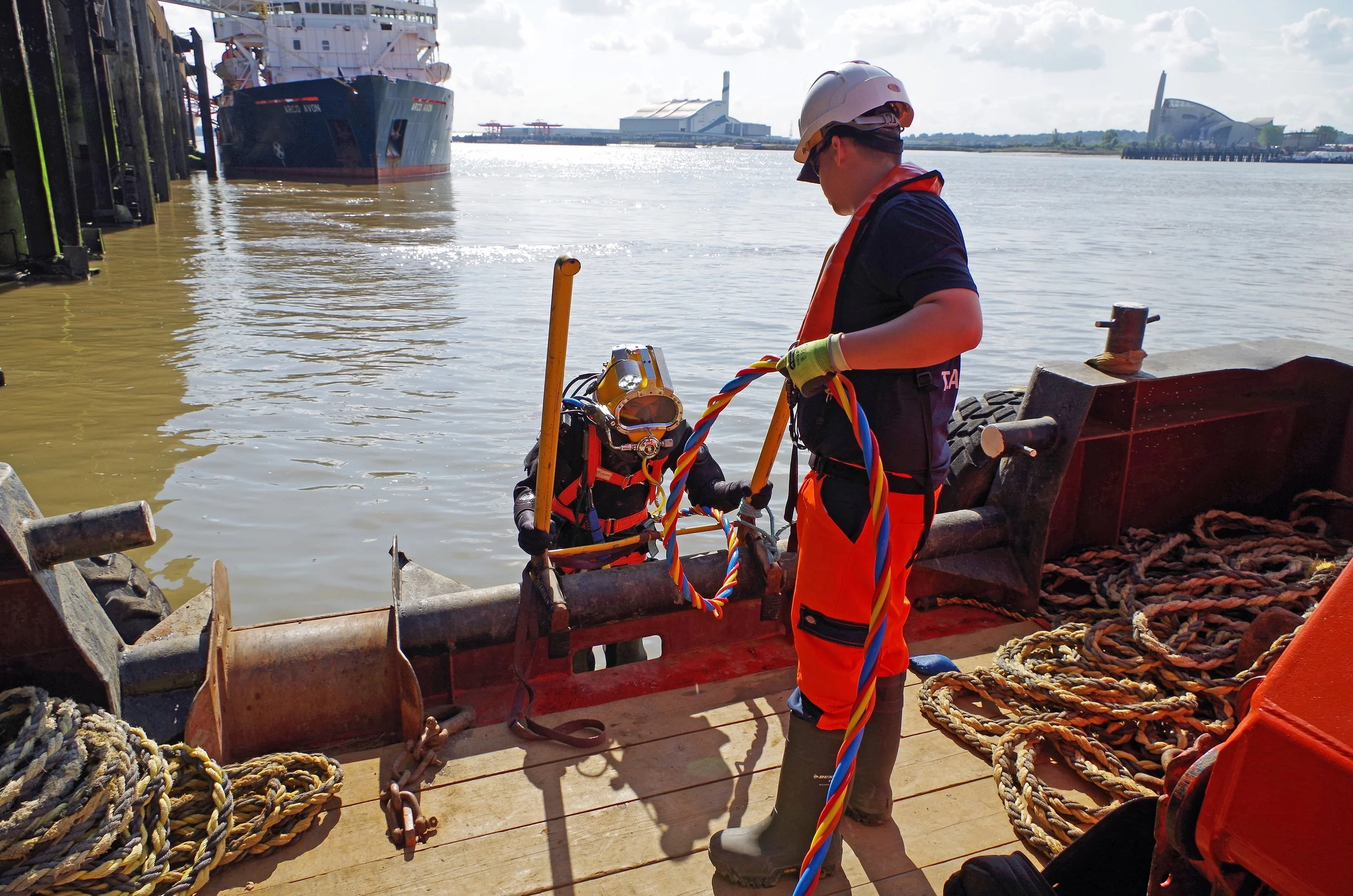 Two workers in safety gear, including helmets and gloves, working on a boat near the water. One is kneeling in the water, and the other is standing on the boat, handling ropes and equipment. Ships and a distant building are visible in the background.