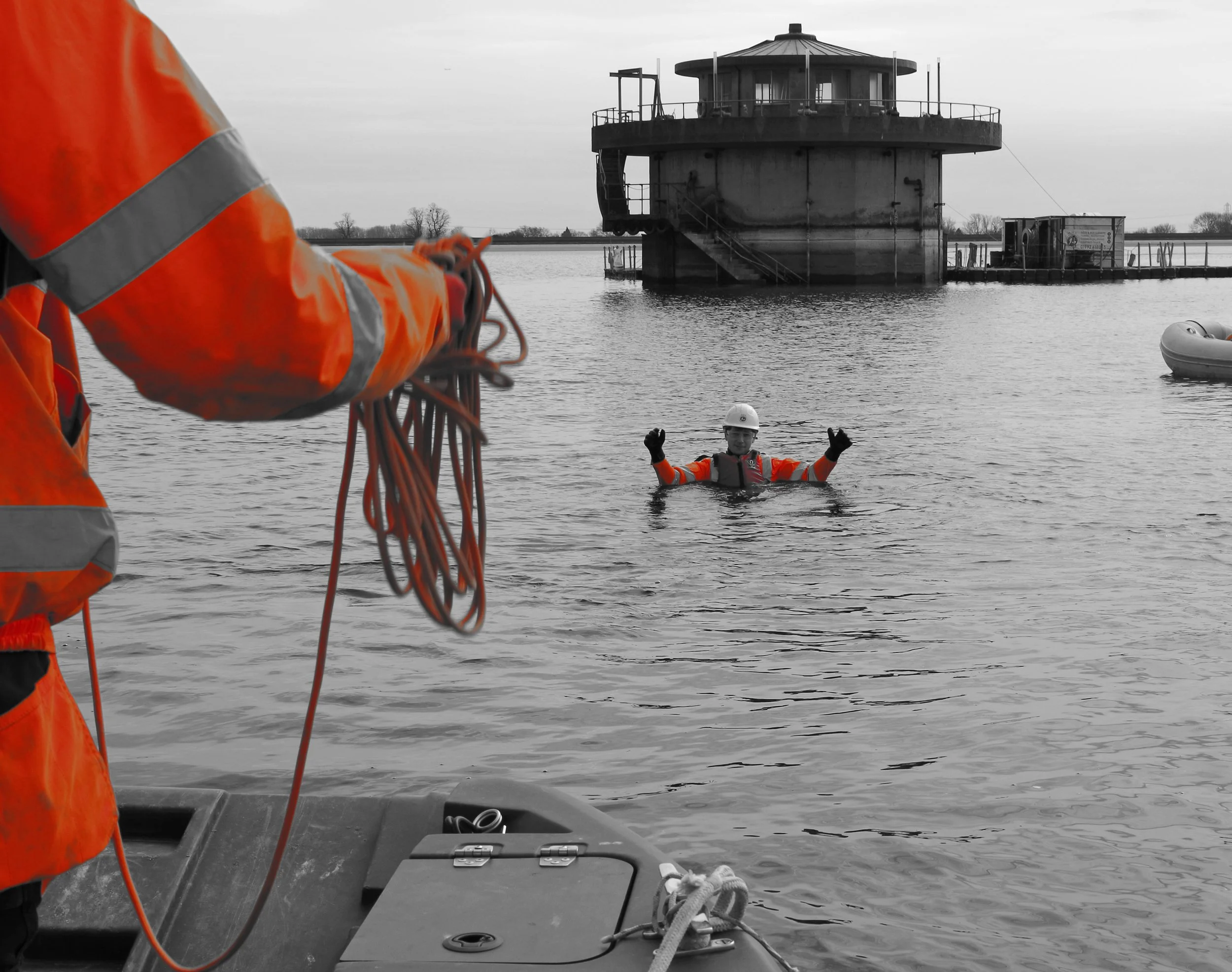 A rescue worker in orange safety gear and another person in a life jacket and helmet in the water near a docked structure, with a boat in the foreground and a gray sky.