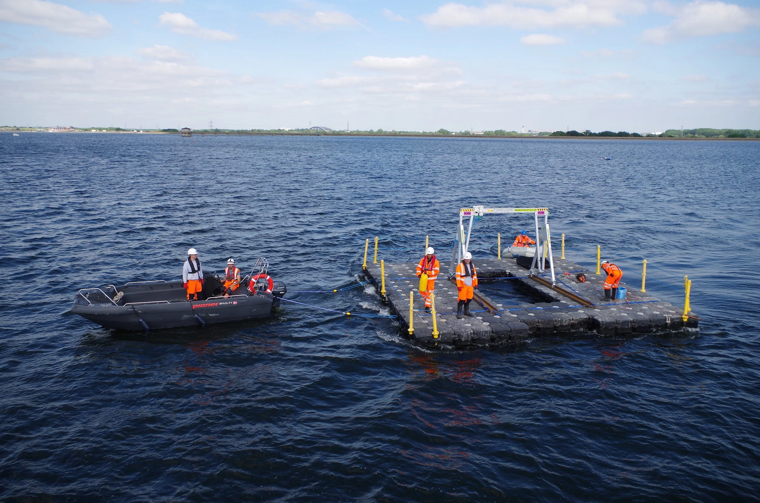 Workers in orange safety gear and helmets on a floating platform with a small boat nearby, in a large body of water under a partly cloudy sky.