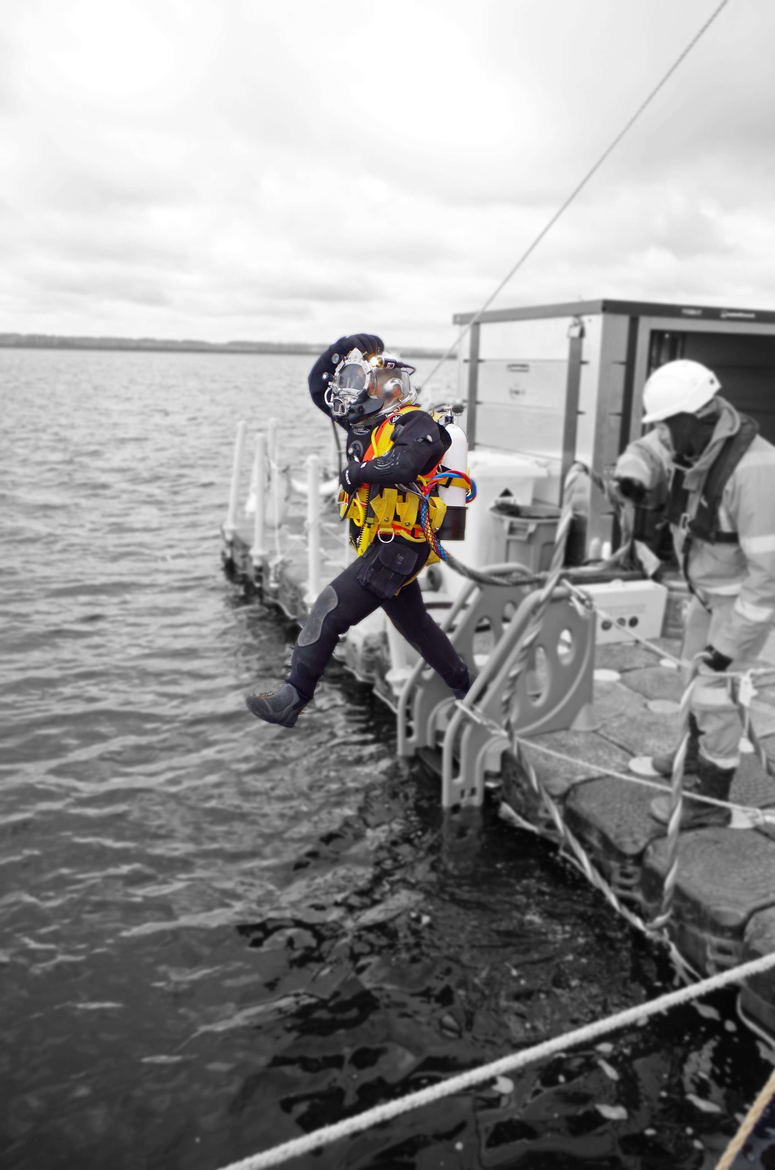 A diver in full gear, including a helmet, exiting a boat into the water. The background and the person are in color, while the rest of the image is in black and white.
