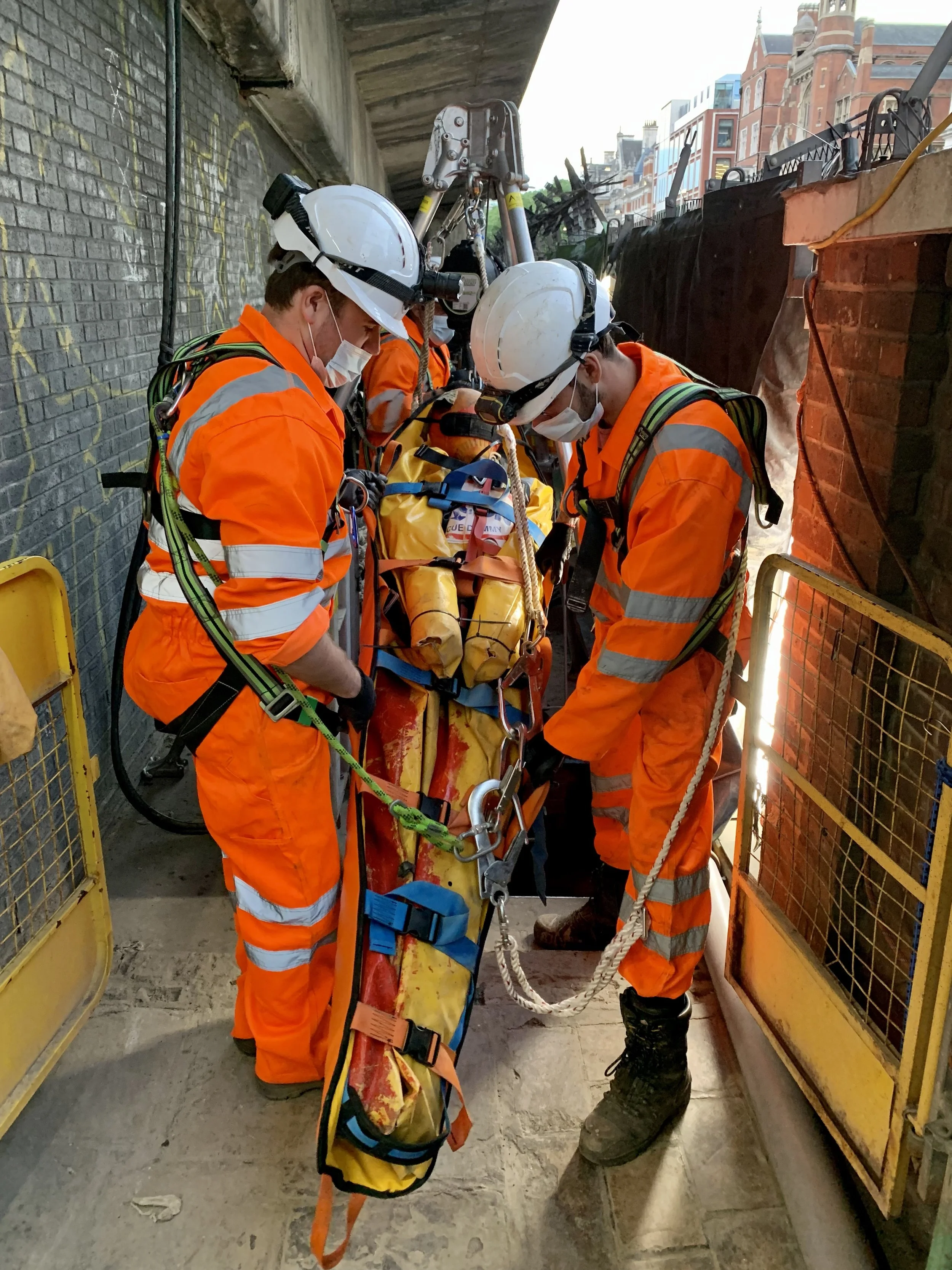 Three construction workers in orange safety uniforms and white helmets are gathered around a person on a stretcher, preparing to lift a rescue dummy for a confined space drill. 