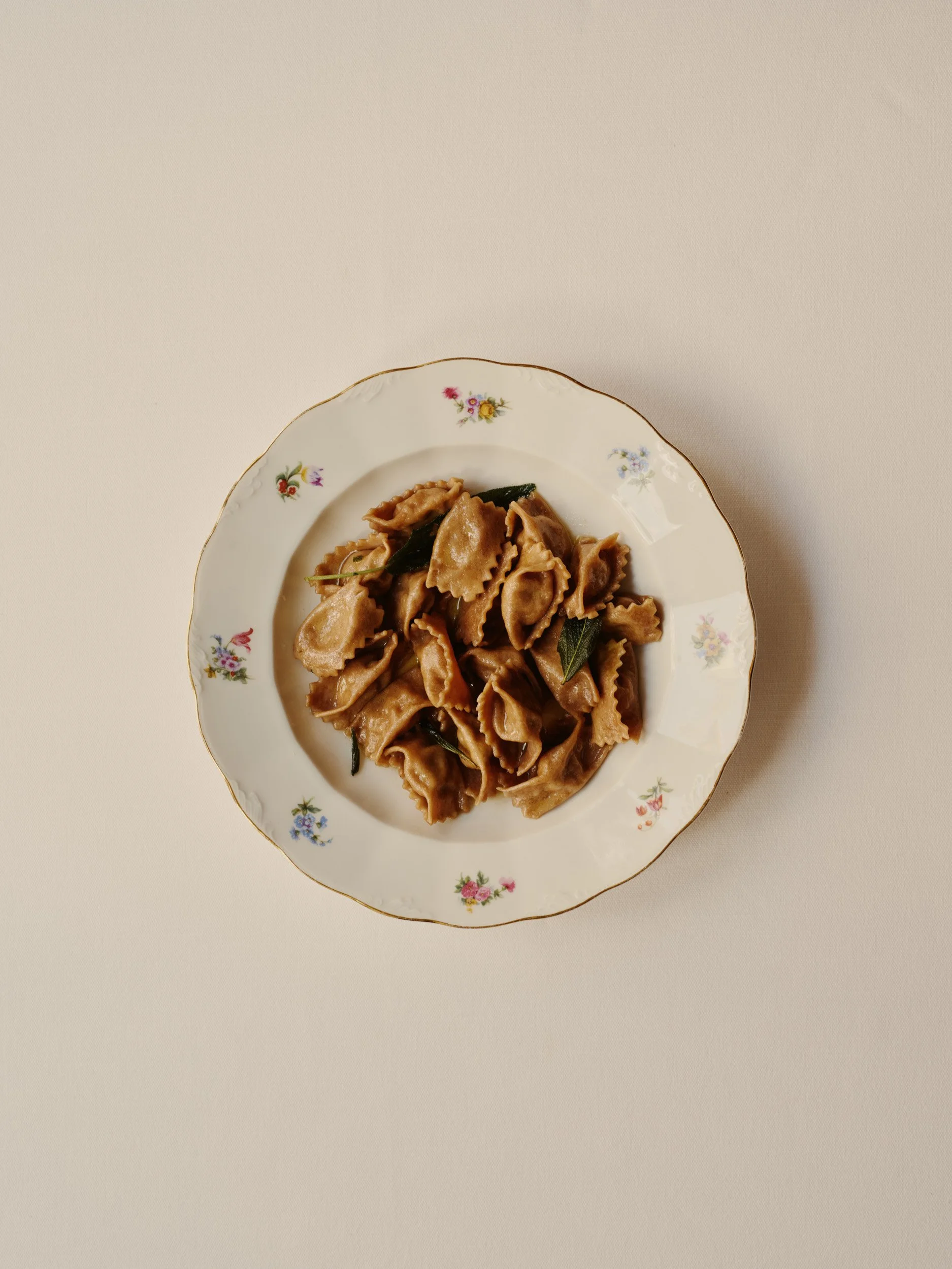 A white porcelain plate with a floral pattern around the rim, filled with cooked bowtie-shaped pasta and garnished with green herbs.