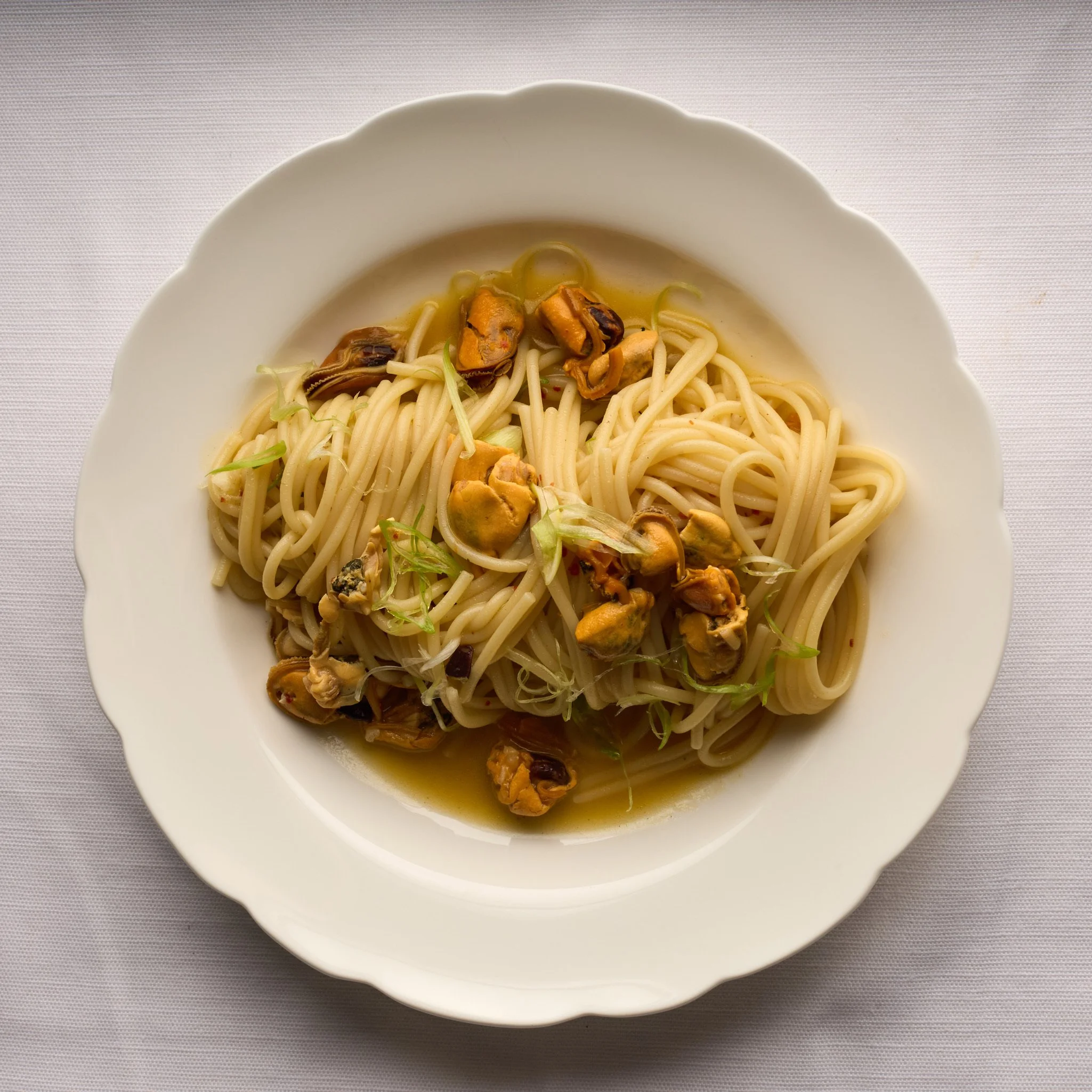 Plate of cooked pasta with shellfish and green garnish, in broth on a white dish.
