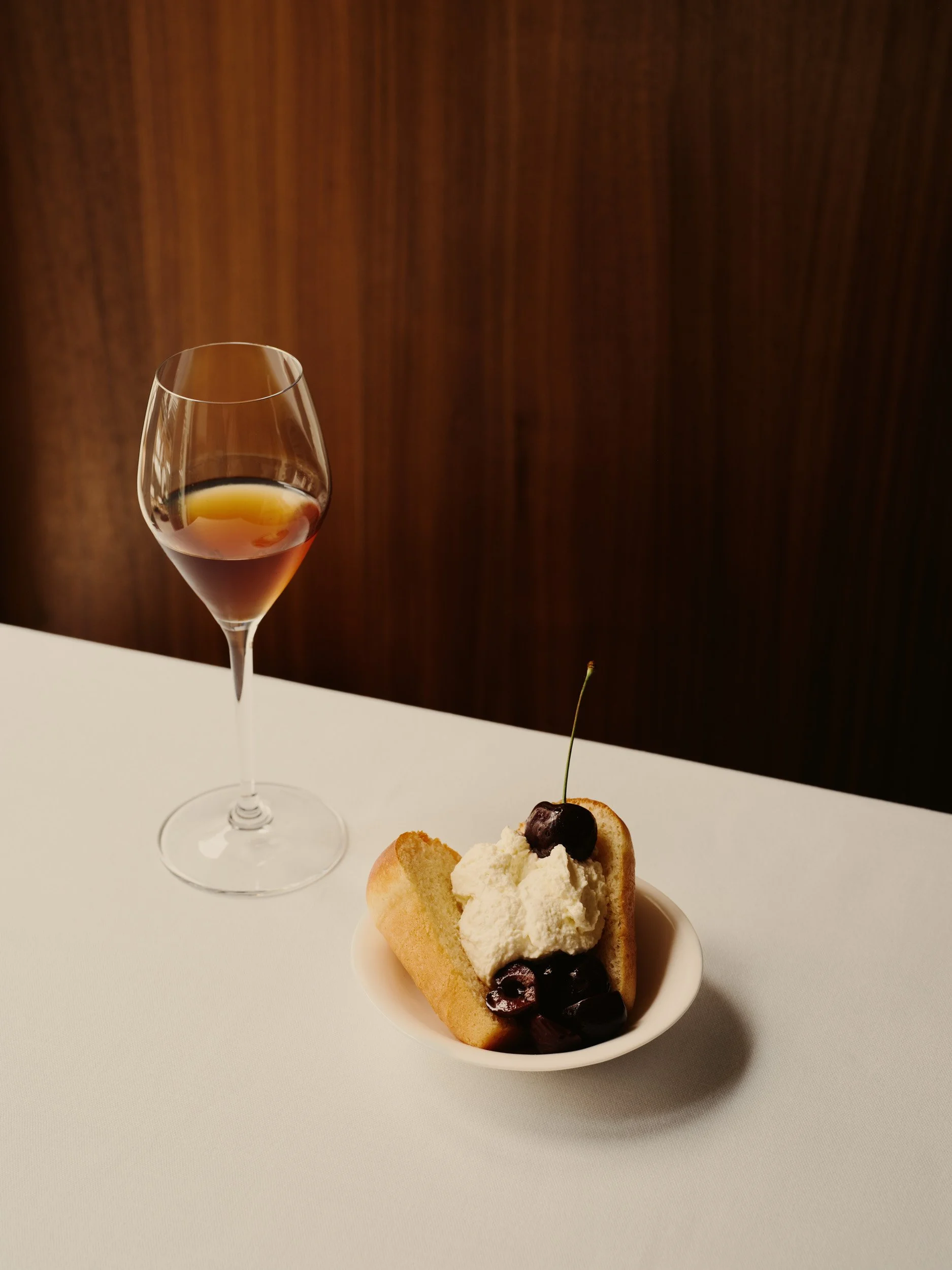 A glass of red wine and a small dish with a cannoli topped with whipped cream and cherries, on a white table with a wooden background.