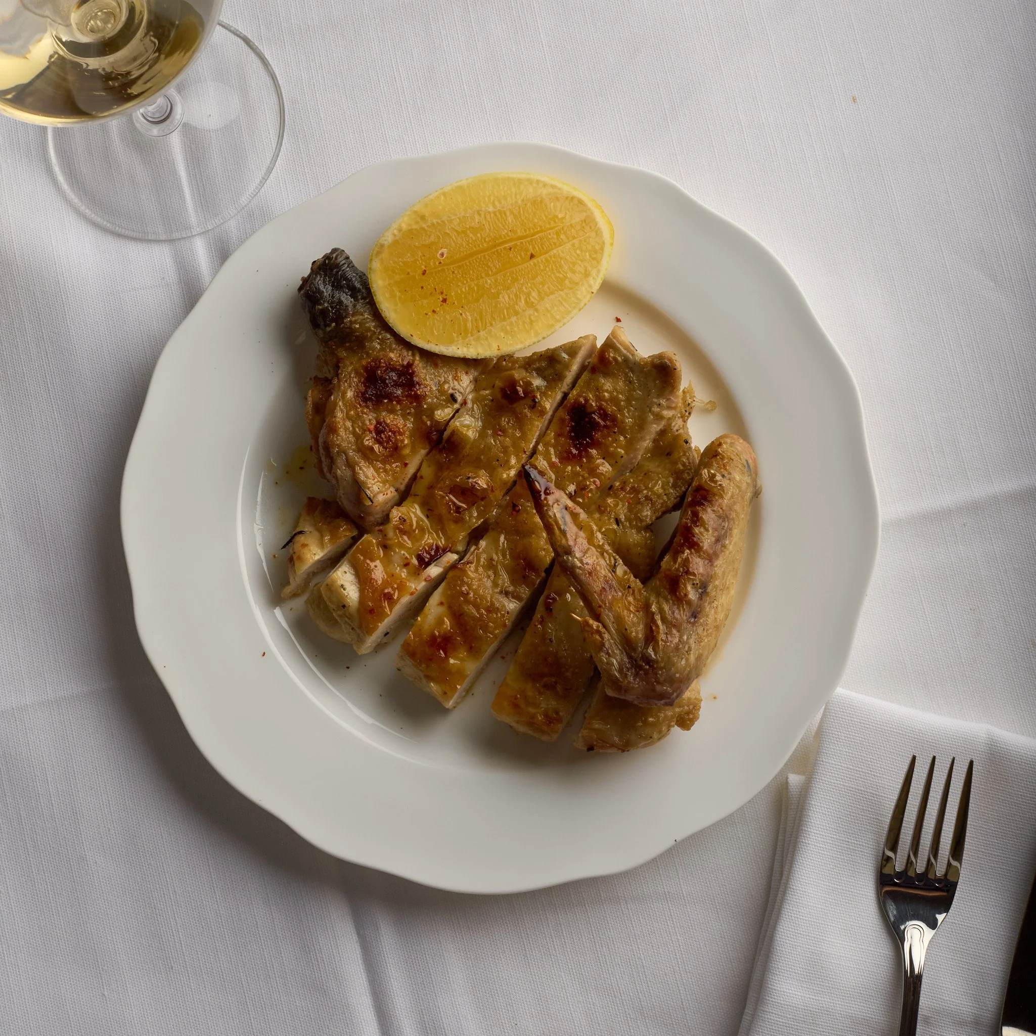 Grilled chicken served with lemon wedge on a white plate, with a glass of white wine nearby on a white tablecloth.