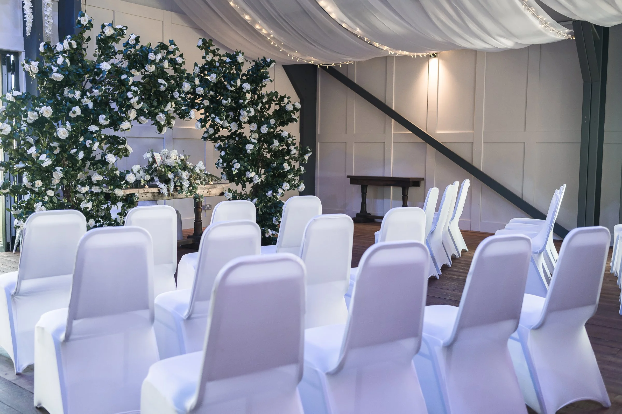Indoor wedding ceremony setup with white chairs arranged in rows facing a floral backdrop with white and green flowers. Draped fabric and string lights decorate the ceiling.