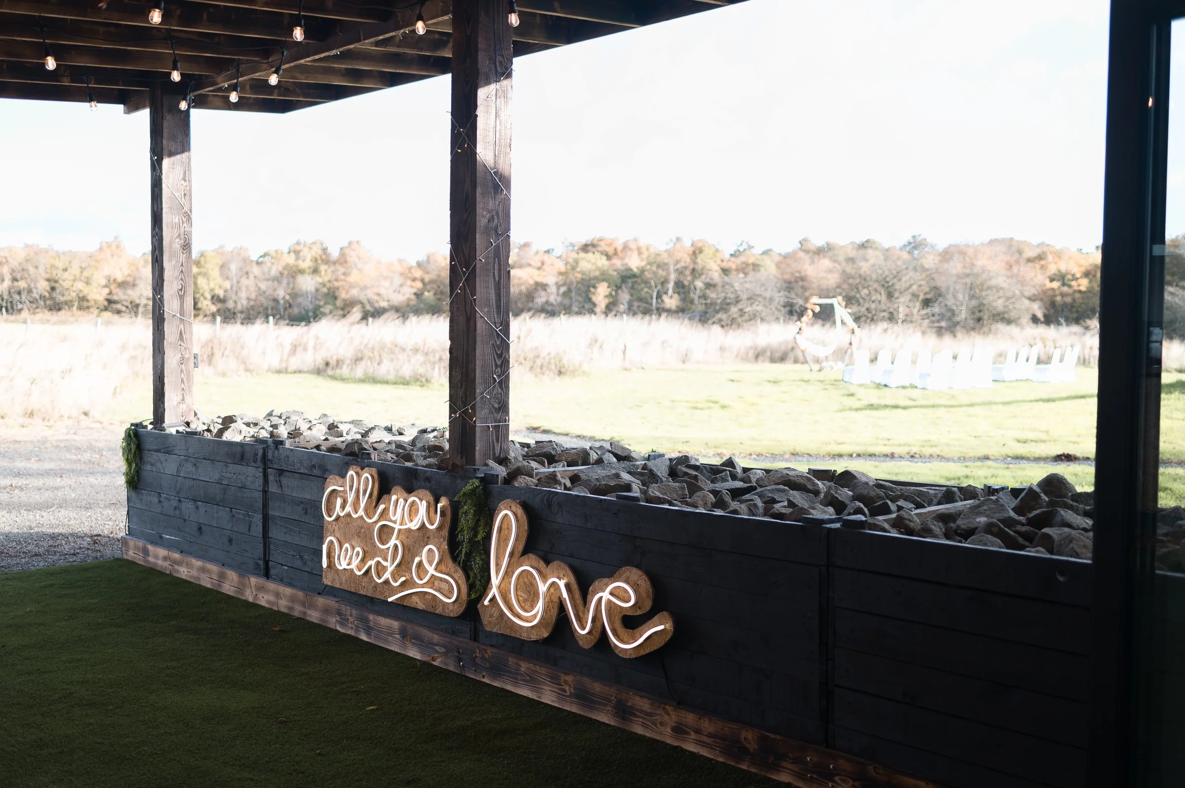 Rustic outdoor wedding decor with a sign that says 'all you need is love' mounted on a dark wooden planter box filled with rocks, overlooking a grassy field with chairs and a swing set in the distance.