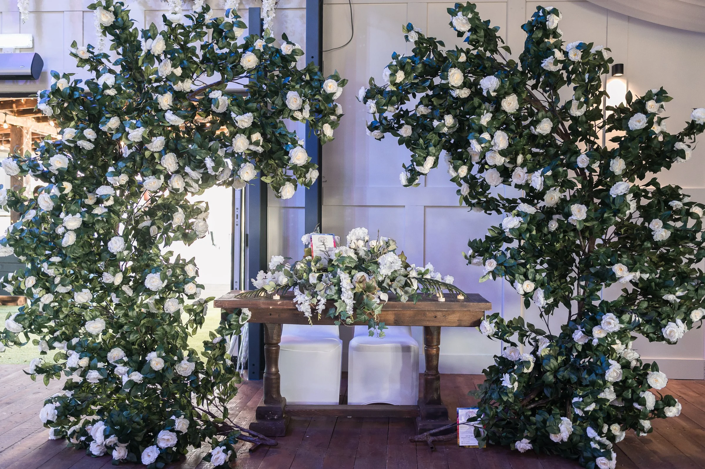 Decorative floral setup with large white flowers and green leaves, surrounding a wooden table with white chairs inside a well-lit room.