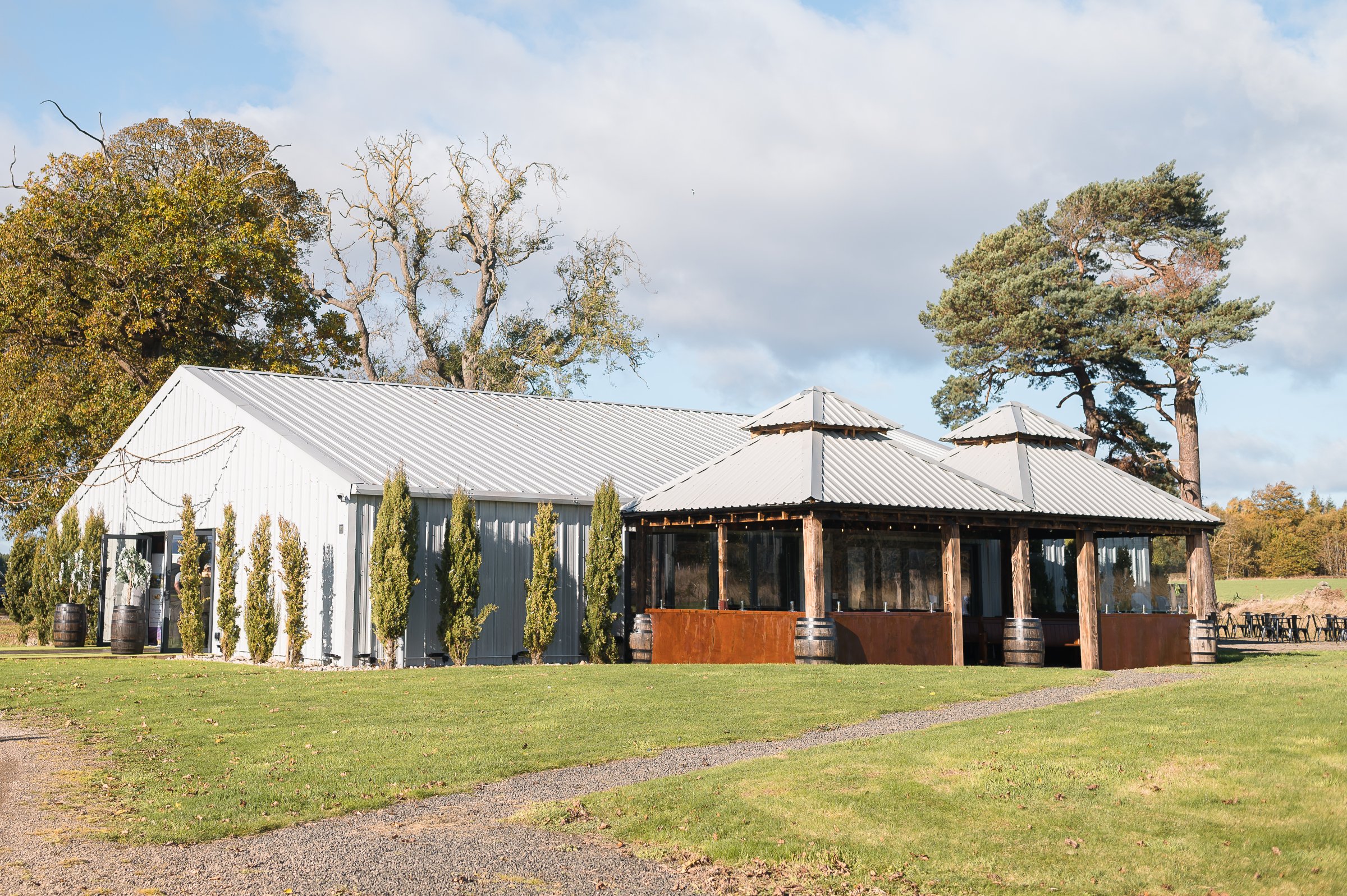 A rustic outdoor pavilion with a metal roof, wooden beams, and glass walls set in a grassy area surrounded by trees.