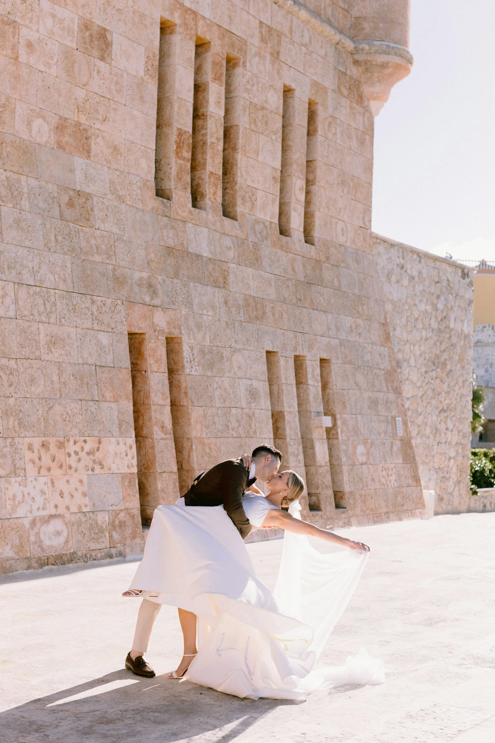 A couple in wedding attire, with the groom dipping the bride for a kiss, standing outdoors in front of a stone building with small rectangular windows.