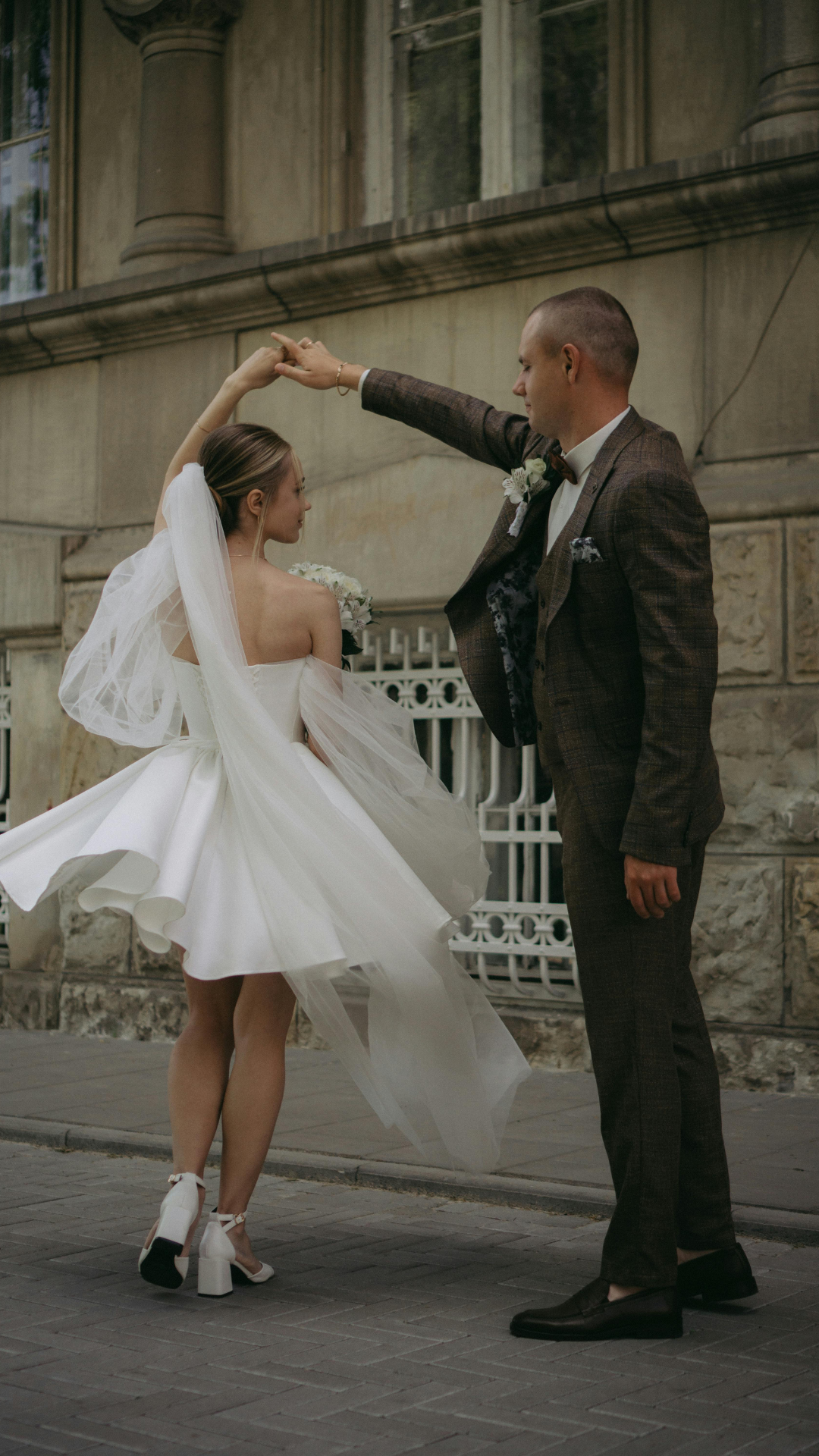 A couple dancing outdoors, the woman in a white wedding dress and the man in a brown checked suit.