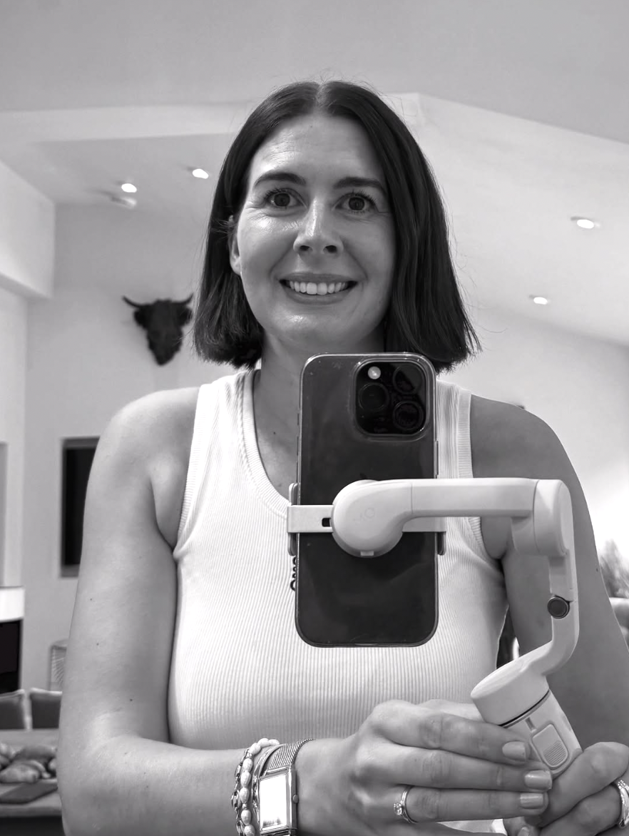 Black and white photo of a woman taking a selfie with a smartphone mounted on a handheld stabilizer. She has shoulder-length dark hair, is smiling, and wearing a sleeveless top. In the background, a wall with a mounted animal head is visible.