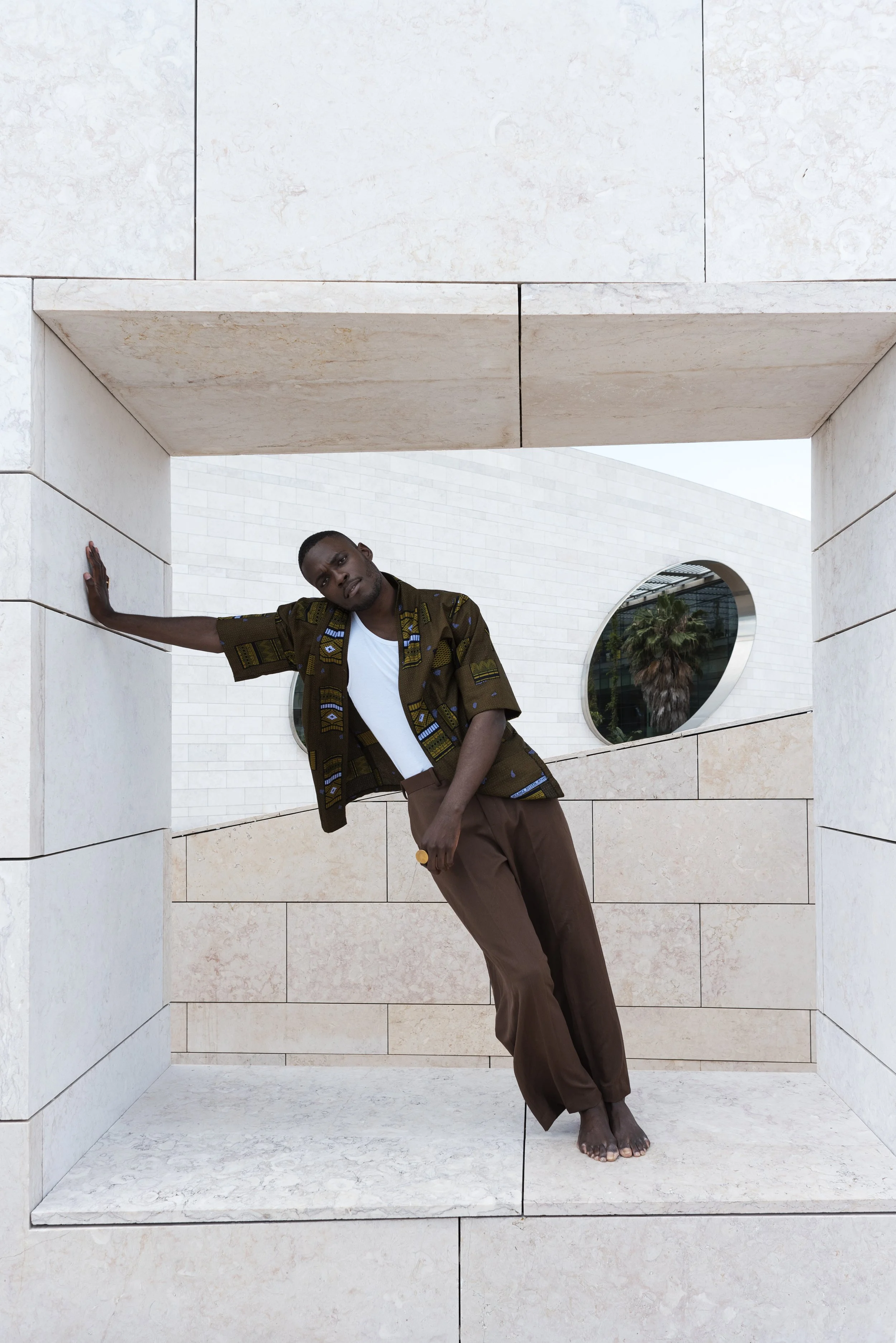 A man standing barefoot in a modern architectural space with light-colored stone walls and a circular window showing palm trees outside.