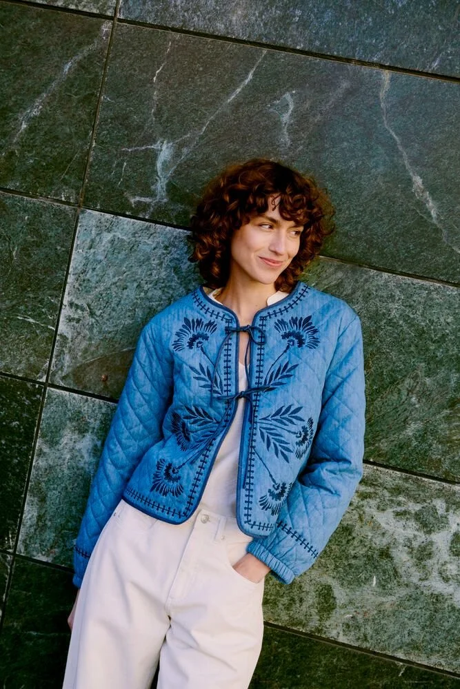 Two women standing outside between white buildings with a clear blue sky in the background. They are dressed in casual clothing, with the woman on the right wearing a leather jacket and the woman on the left wearing a black jacket. Both have shoulder-length hair and serious expressions.
