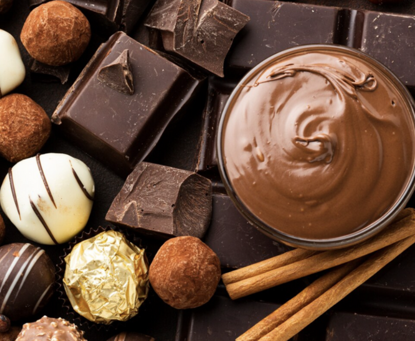 A variety of chocolates, including truffles, chocolate squares, a cup of melted chocolate, and cinnamon sticks, arranged on a dark surface.