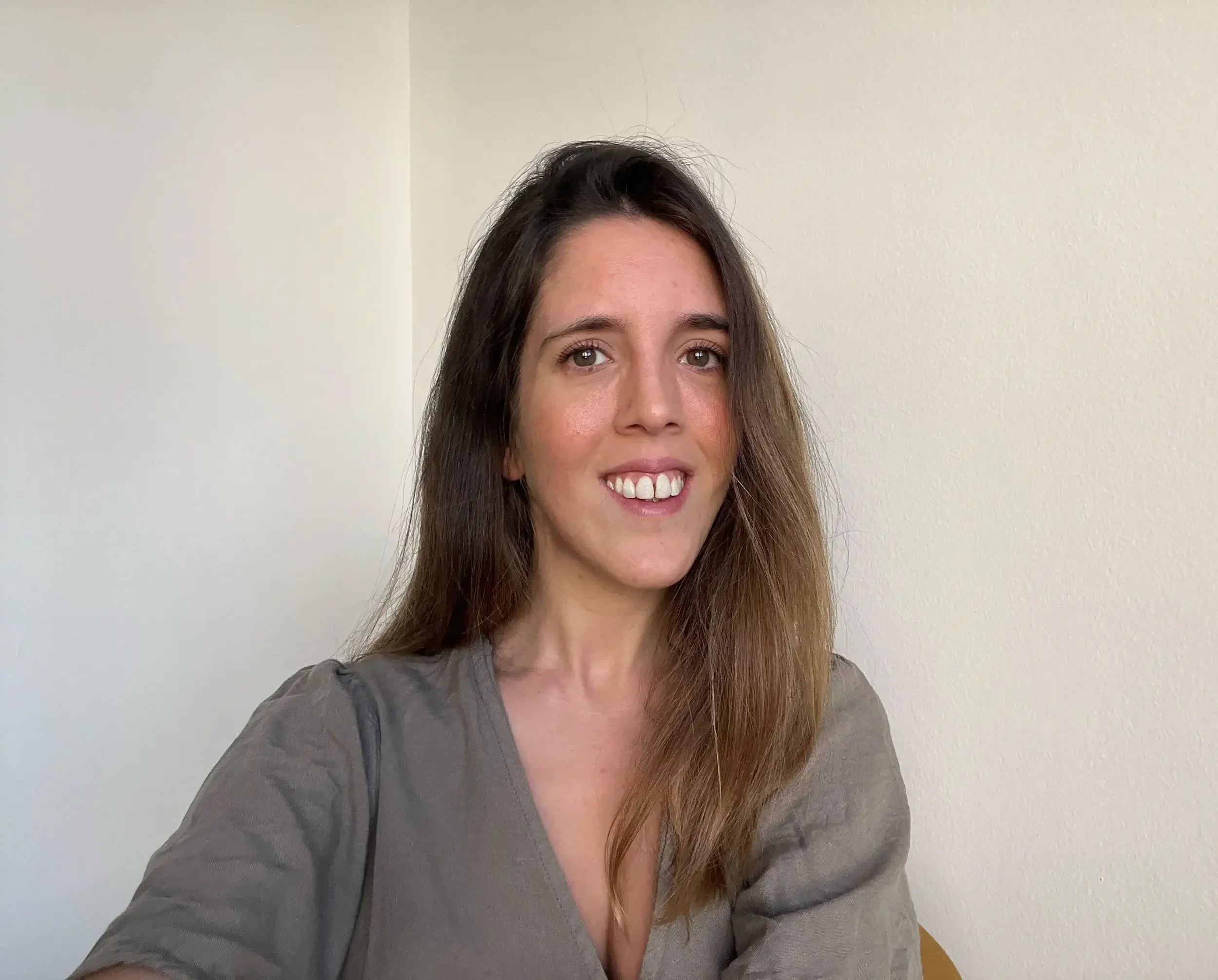Portrait of a woman with brown hair, wearing a grey shirt, standing in front of a white wall smiling
