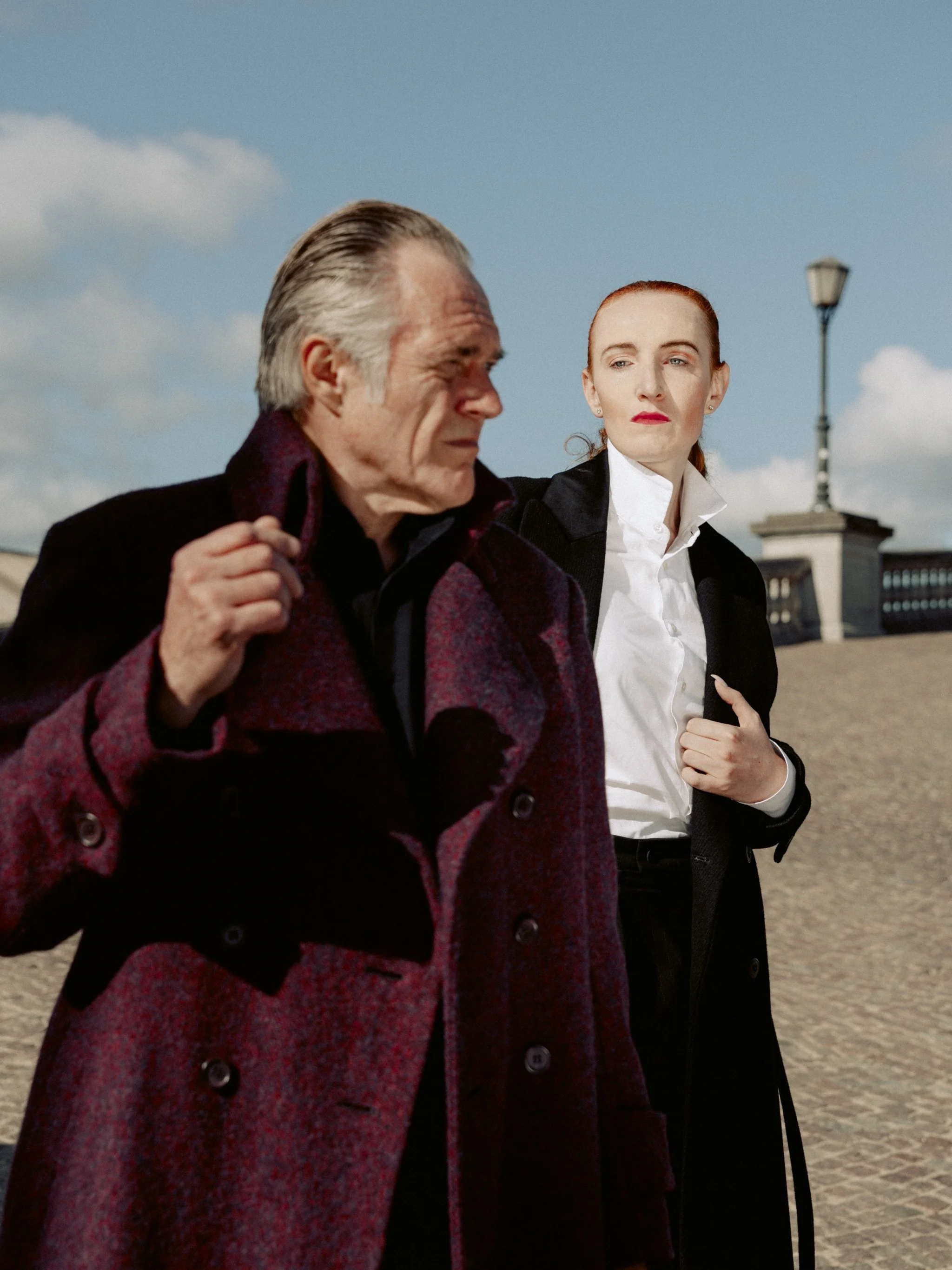 A man with gray hair in a dark maroon coat and a woman with red hair in a black coat and white shirt walking outdoors on a sunny day with clear skies and clouds.