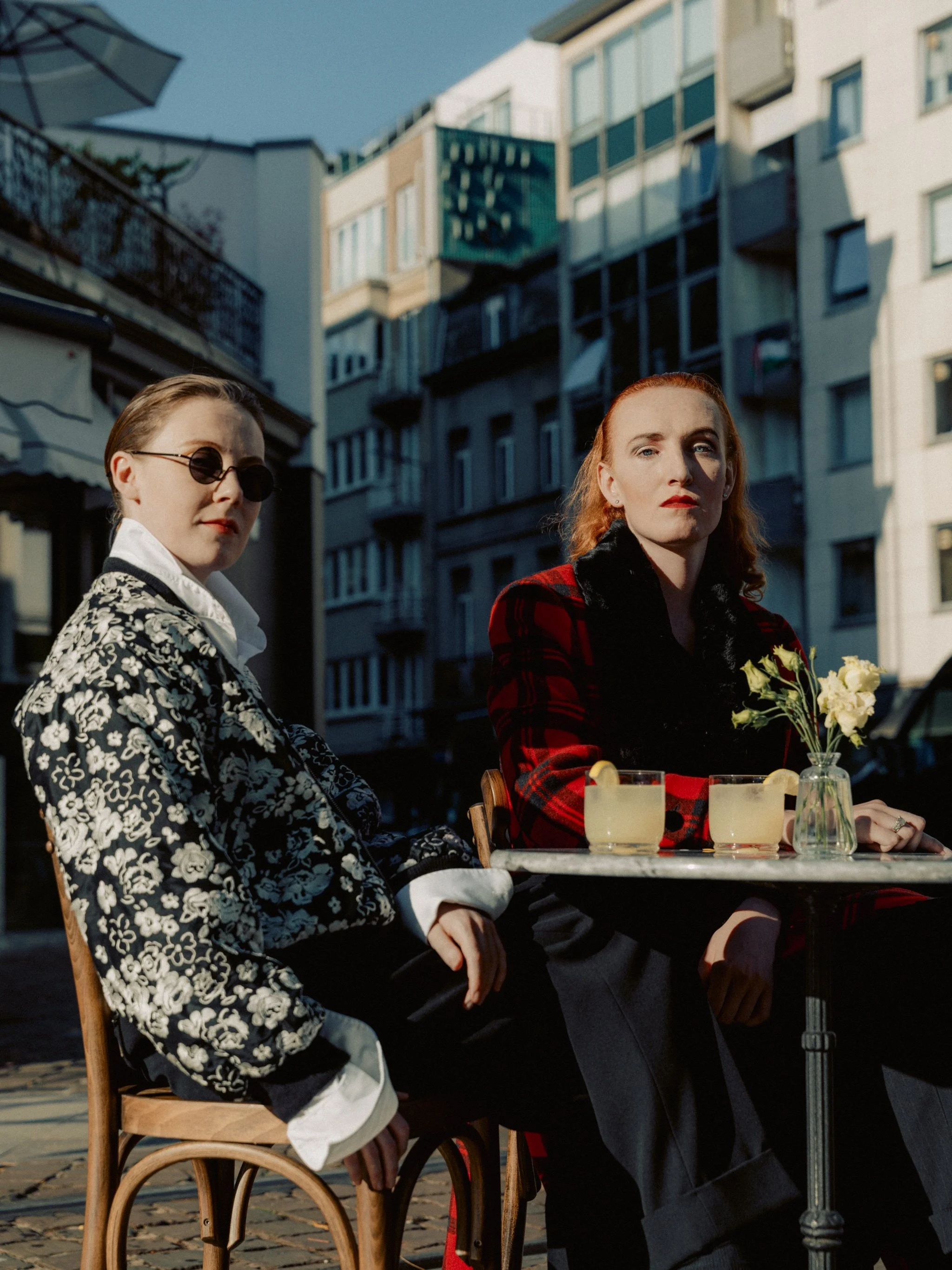 Two women sitting at an outdoor café table with drinks and a small flower vase, in a city street with modern buildings in the background, during sunset.