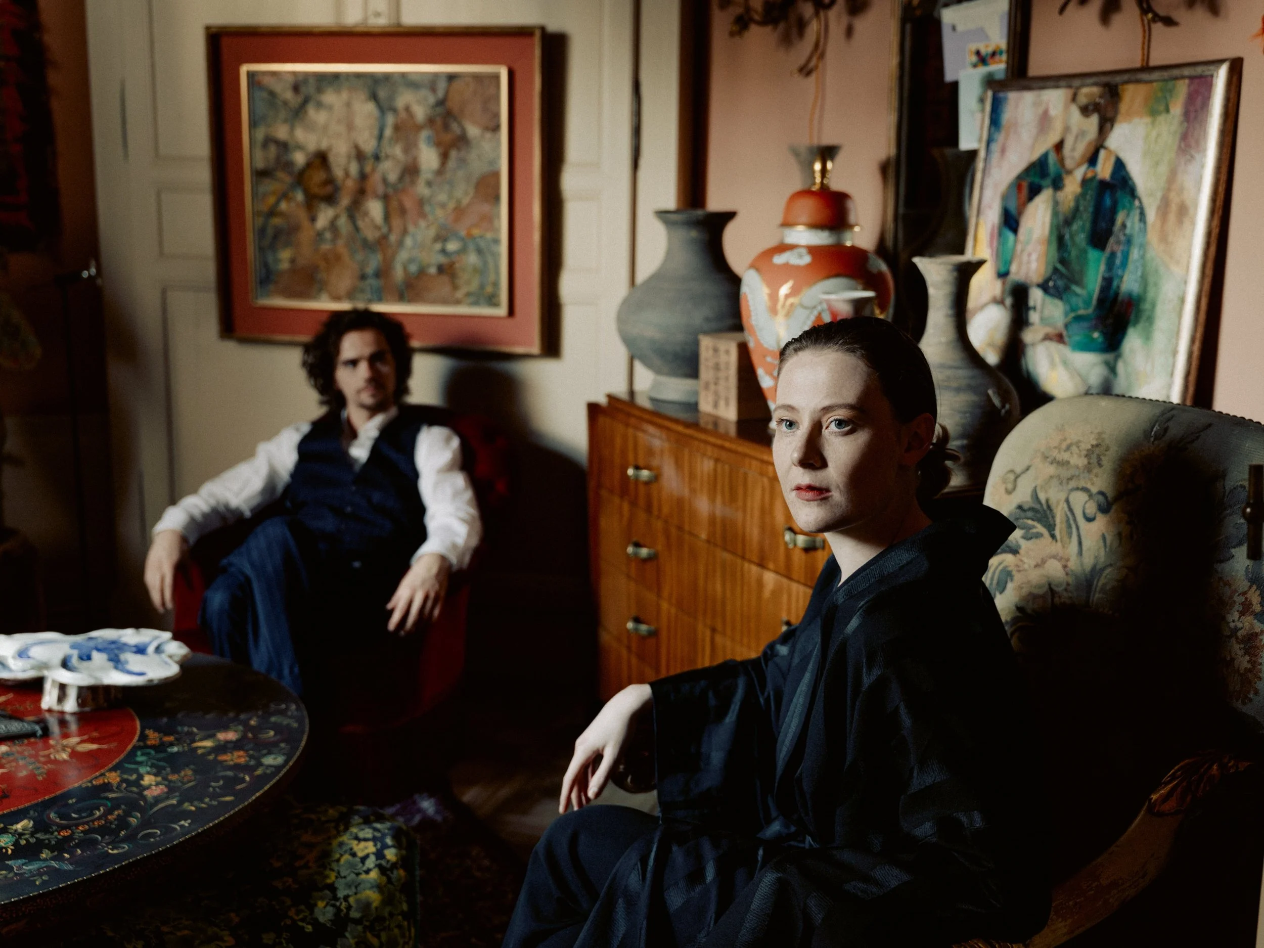 A woman and a man seated in a room with artwork and vases. The woman is in the foreground, looking slightly to the side, wearing a black robe. The man with curly hair and a beard, dressed in a vest and white shirt, is seated in the background, looking towards the woman.