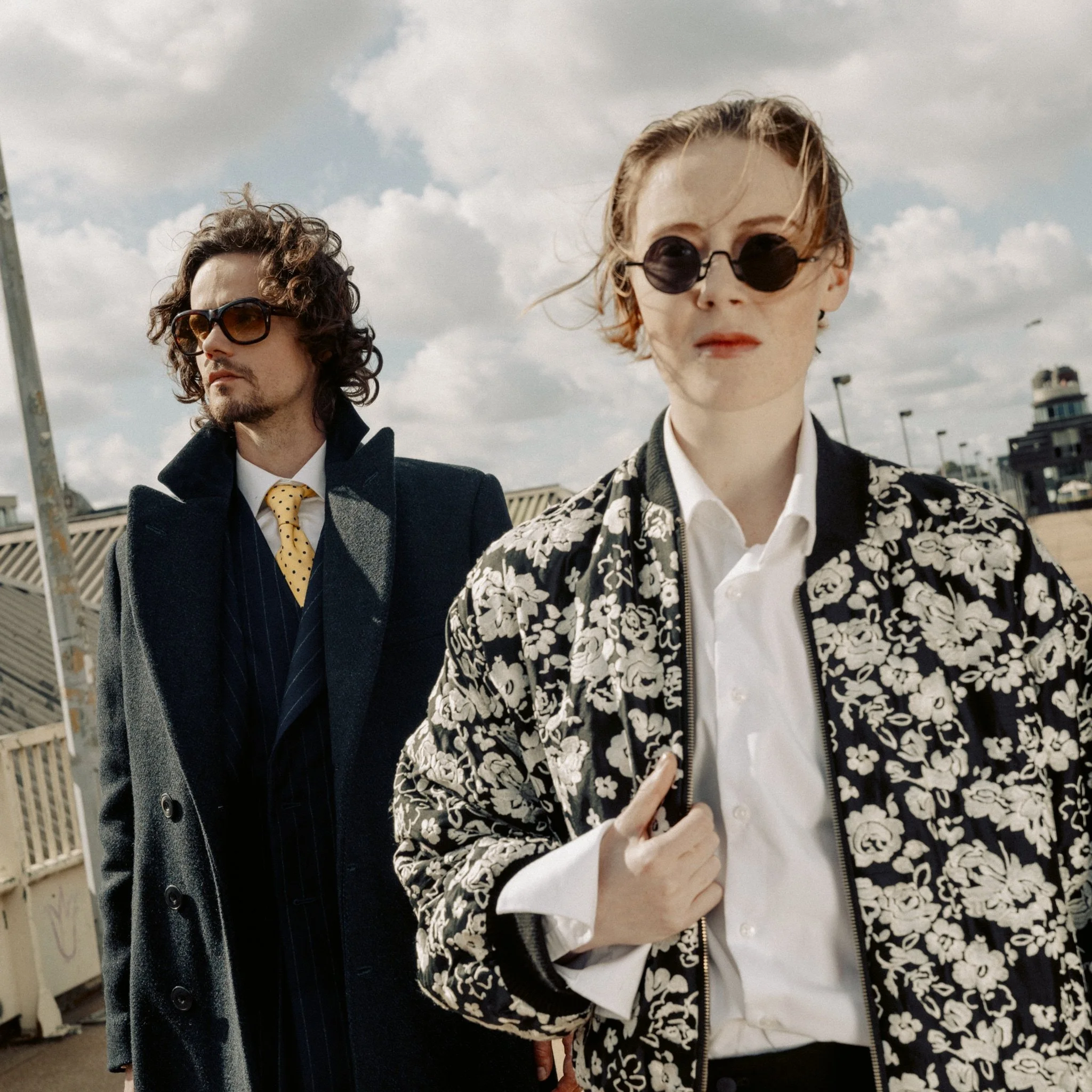 Two stylish individuals outdoors on a cloudy day, both wearing sunglasses and fashionable clothing. The woman in the foreground has short hair, a floral-patterned jacket, and a white shirt. The man in the background has curly hair, a dark overcoat, and a yellow polka-dot tie.