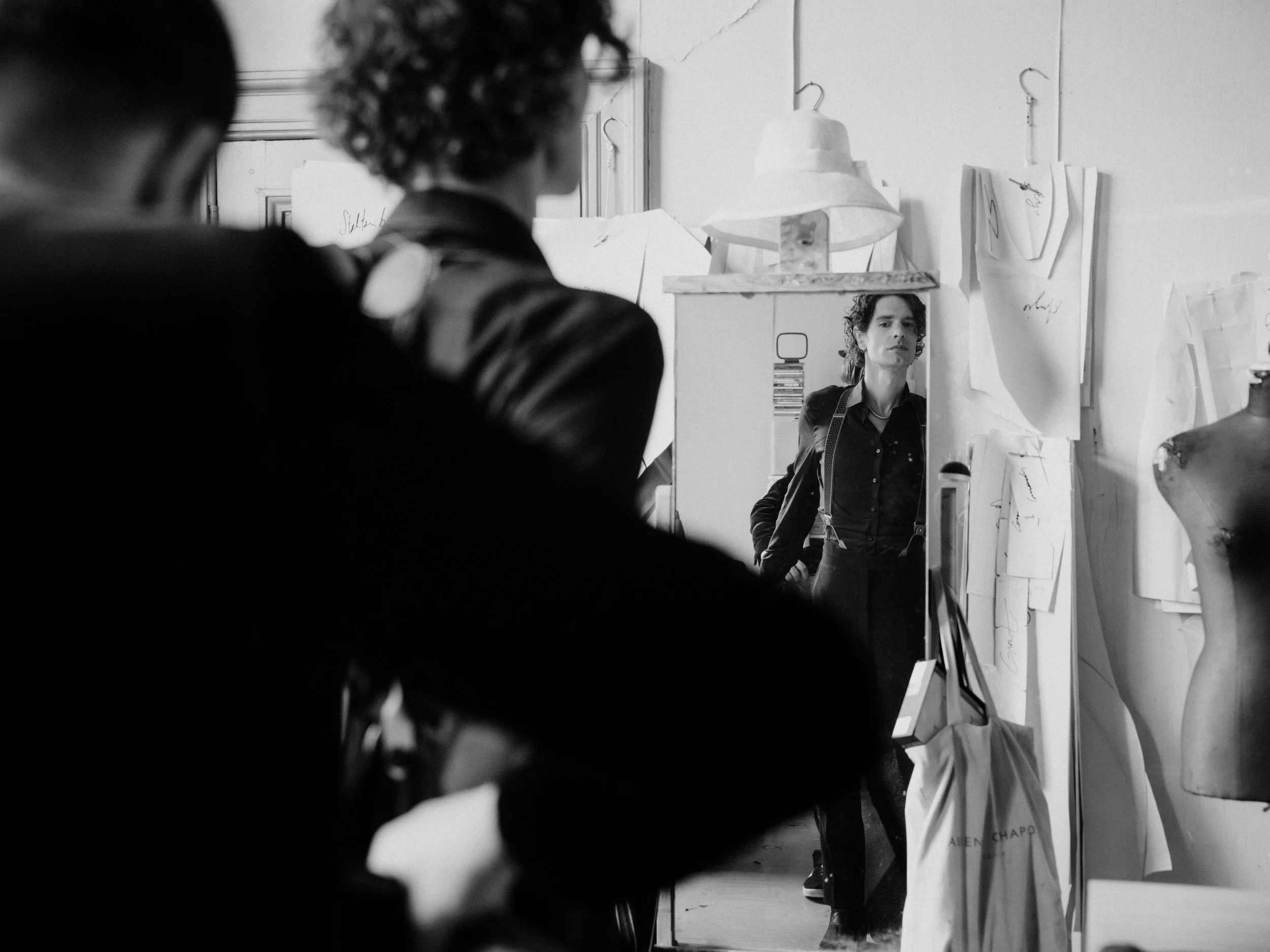 A woman with short hair and suspenders looking at her reflection in a mirror in an artist's studio surrounded by sketches, a mannequin, and art supplies.