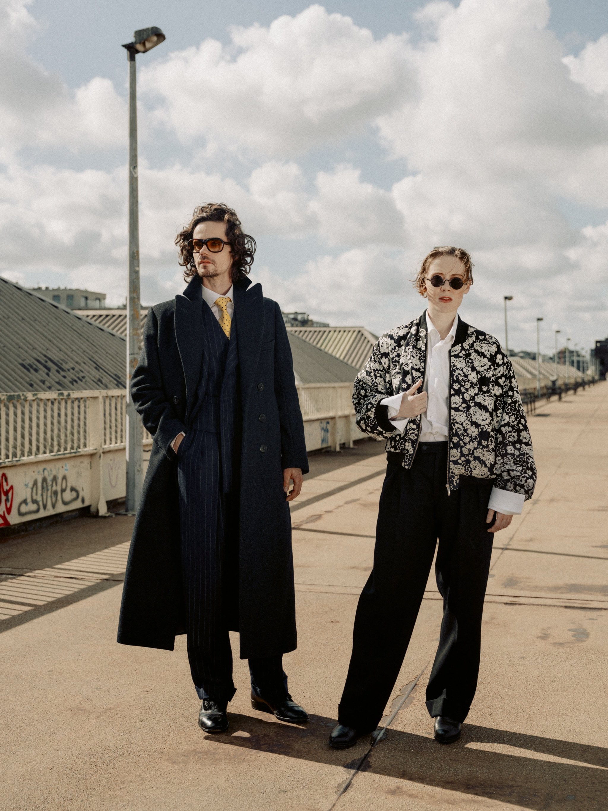 Two people standing on a rooftop in fashionable clothing. The person on the left wears a long black coat, pinstripe pants, and sunglasses, while the person on the right wears a patterned jacket, white shirt, black pants, and sunglasses, with a cloudy sky in the background.