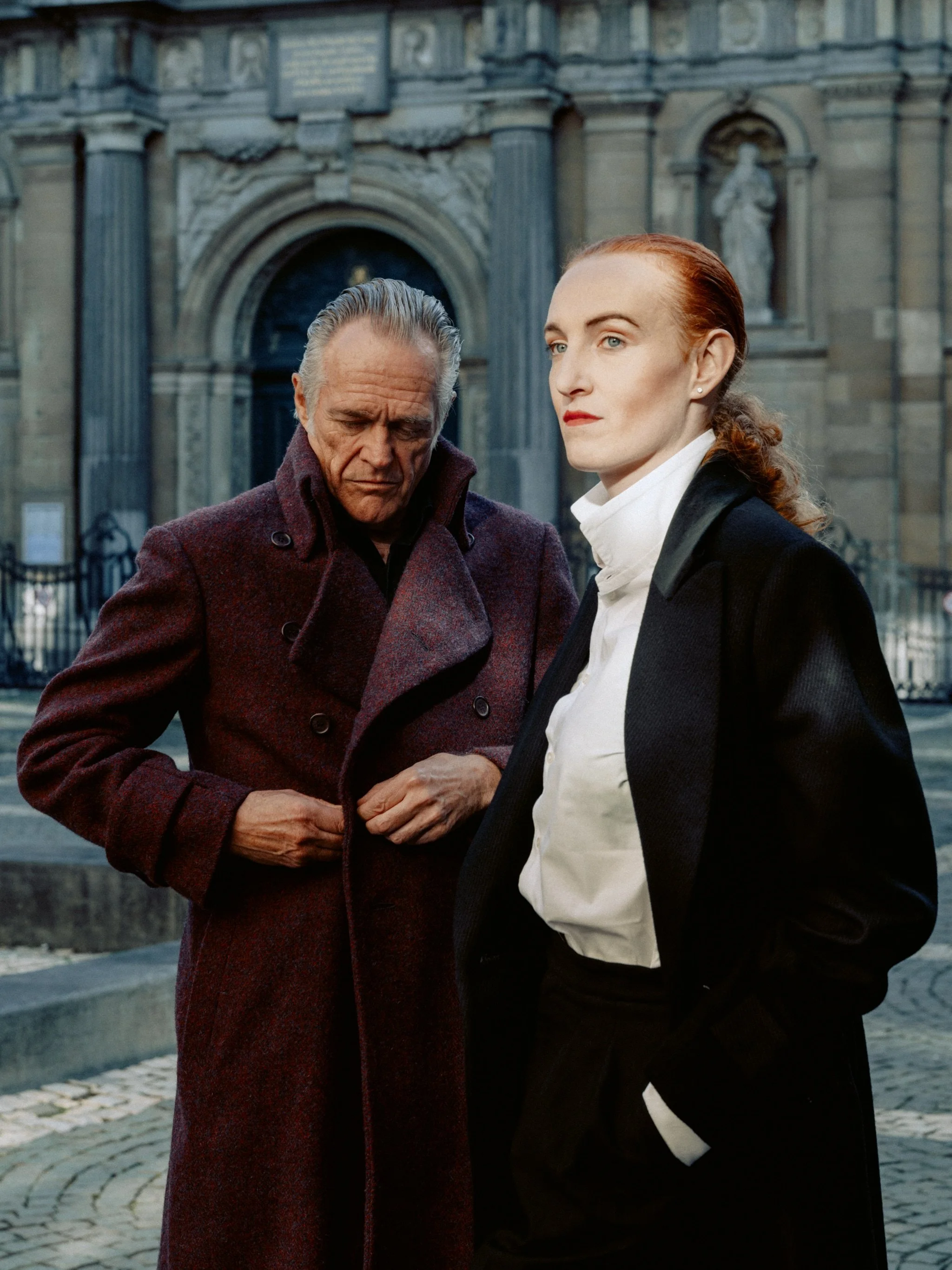 Two people standing in front of a historic building, one man with gray hair in a maroon coat looking down and a woman with red hair in a black coat and white shirt looking ahead.