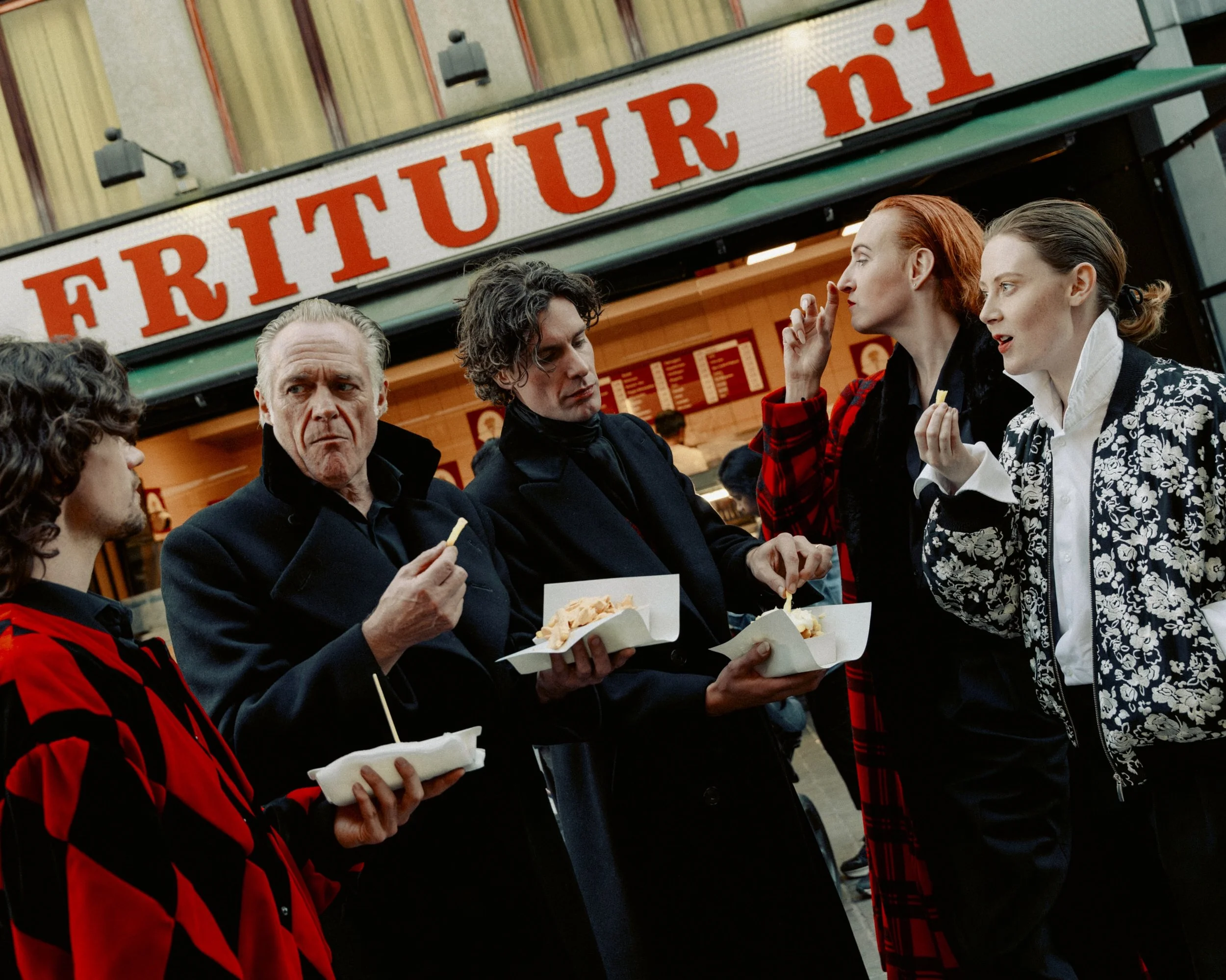 Five people standing outside a food stand named 'Frituur.nl' eating food from paper trays, engaged in conversation.