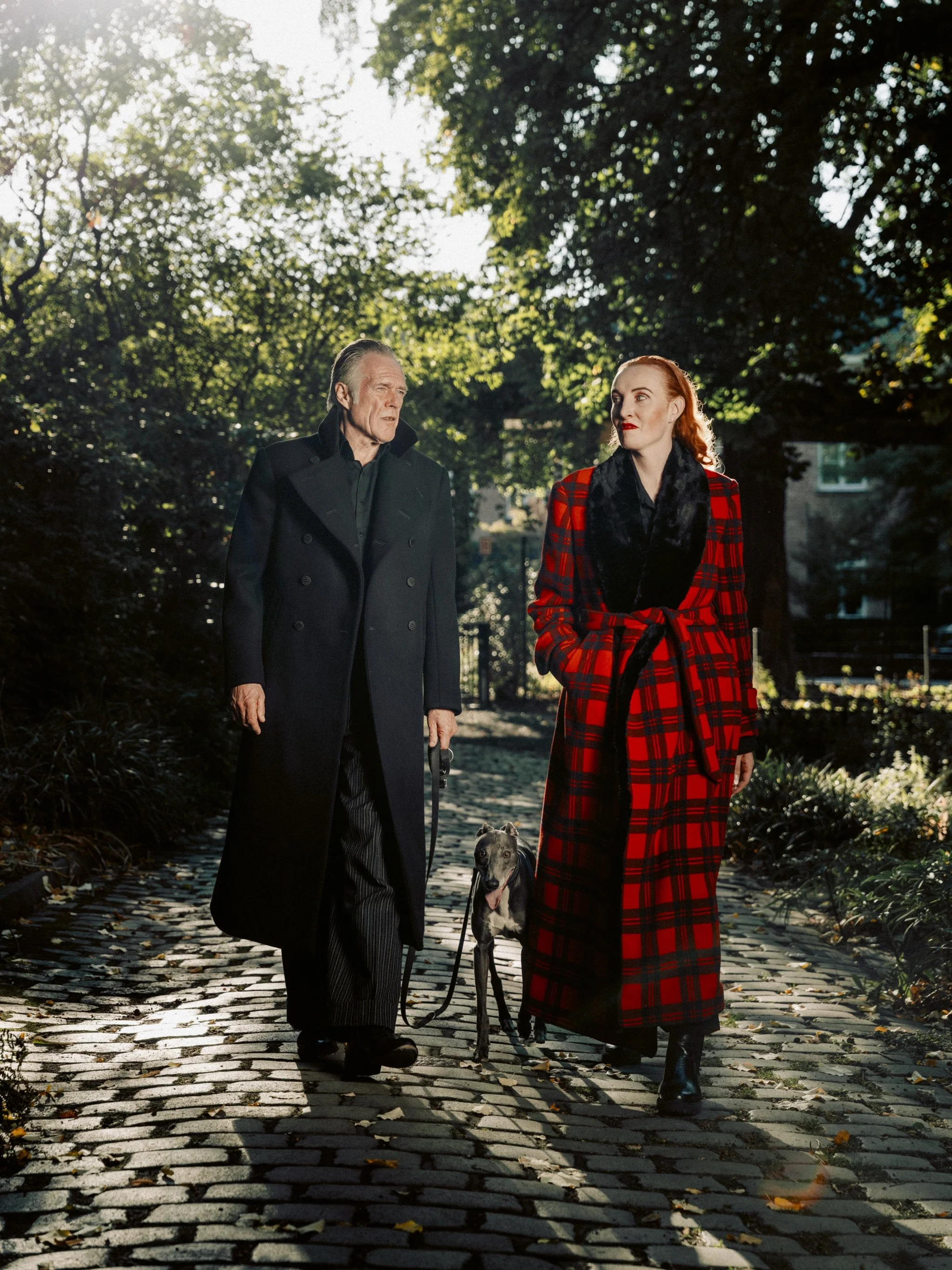 An older man and a young woman walk a dog in a park on a sunny day. The man wears a dark coat, and the woman wears a red and black plaid coat. The background features green trees and sunlight filtering through leaves.