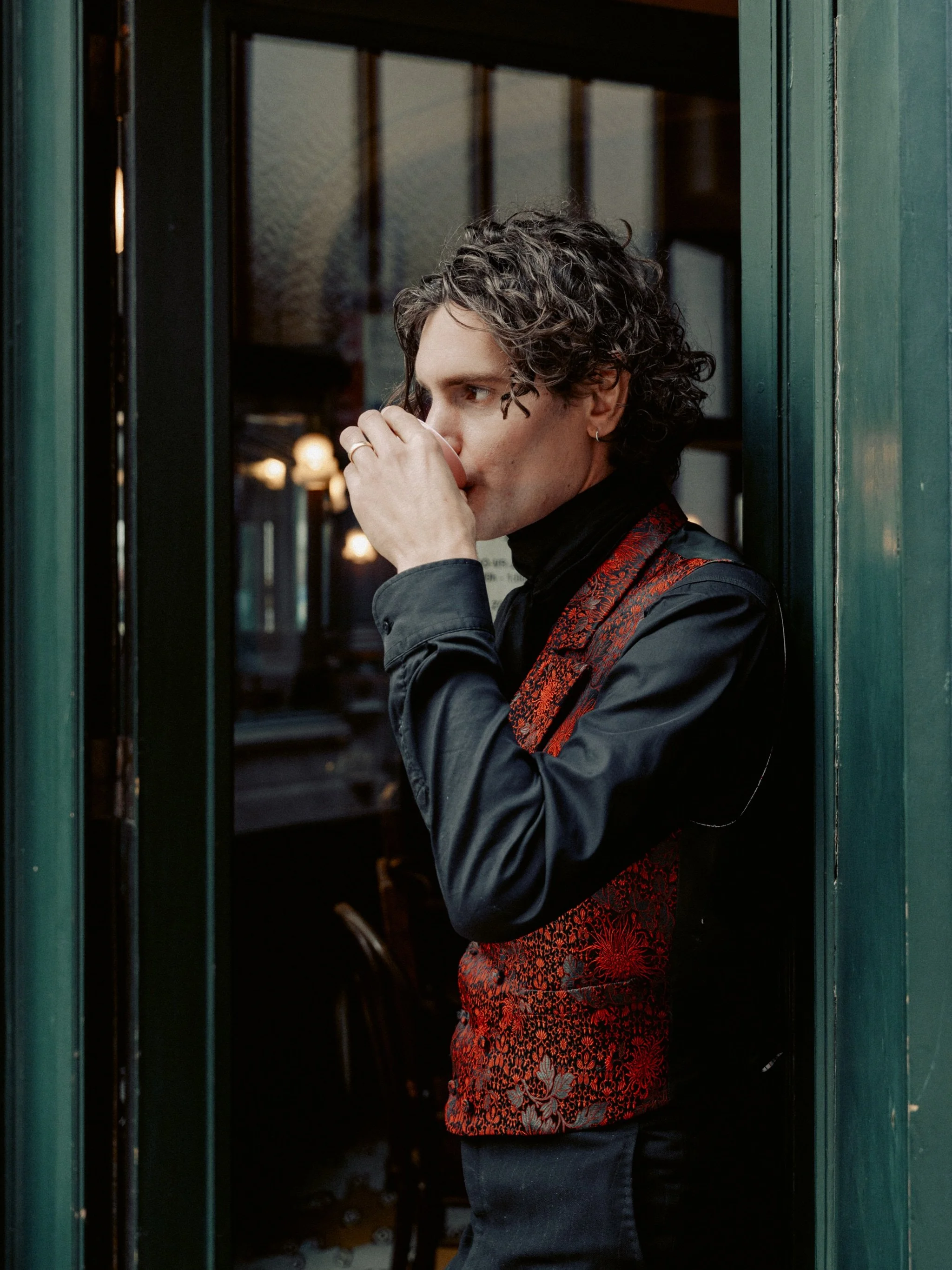 A man with curly dark hair and an earring stands next to a green door or window frame, drinking from a small cup inside a cozy cafe or restaurant.