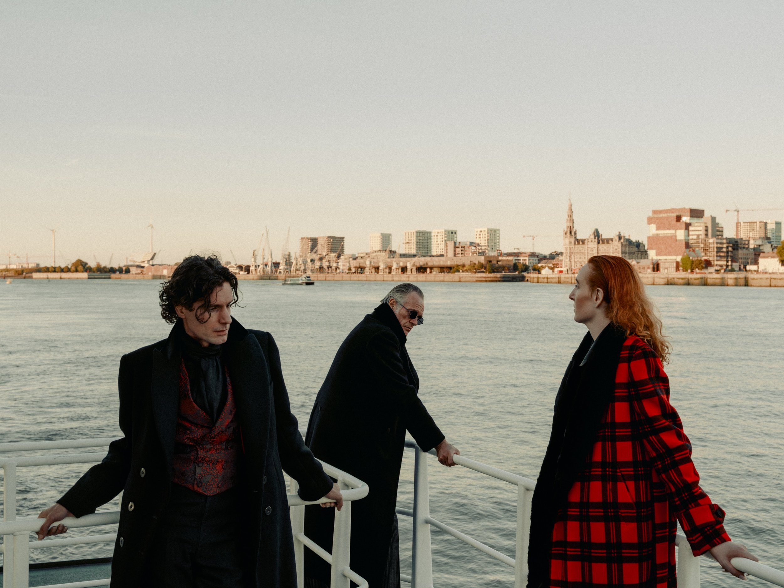 Three people standing on a boat with a city skyline and water in the background. One person with dark hair and a black coat, another person with gray hair and sunglasses, and a woman with red hair and a red plaid coat.