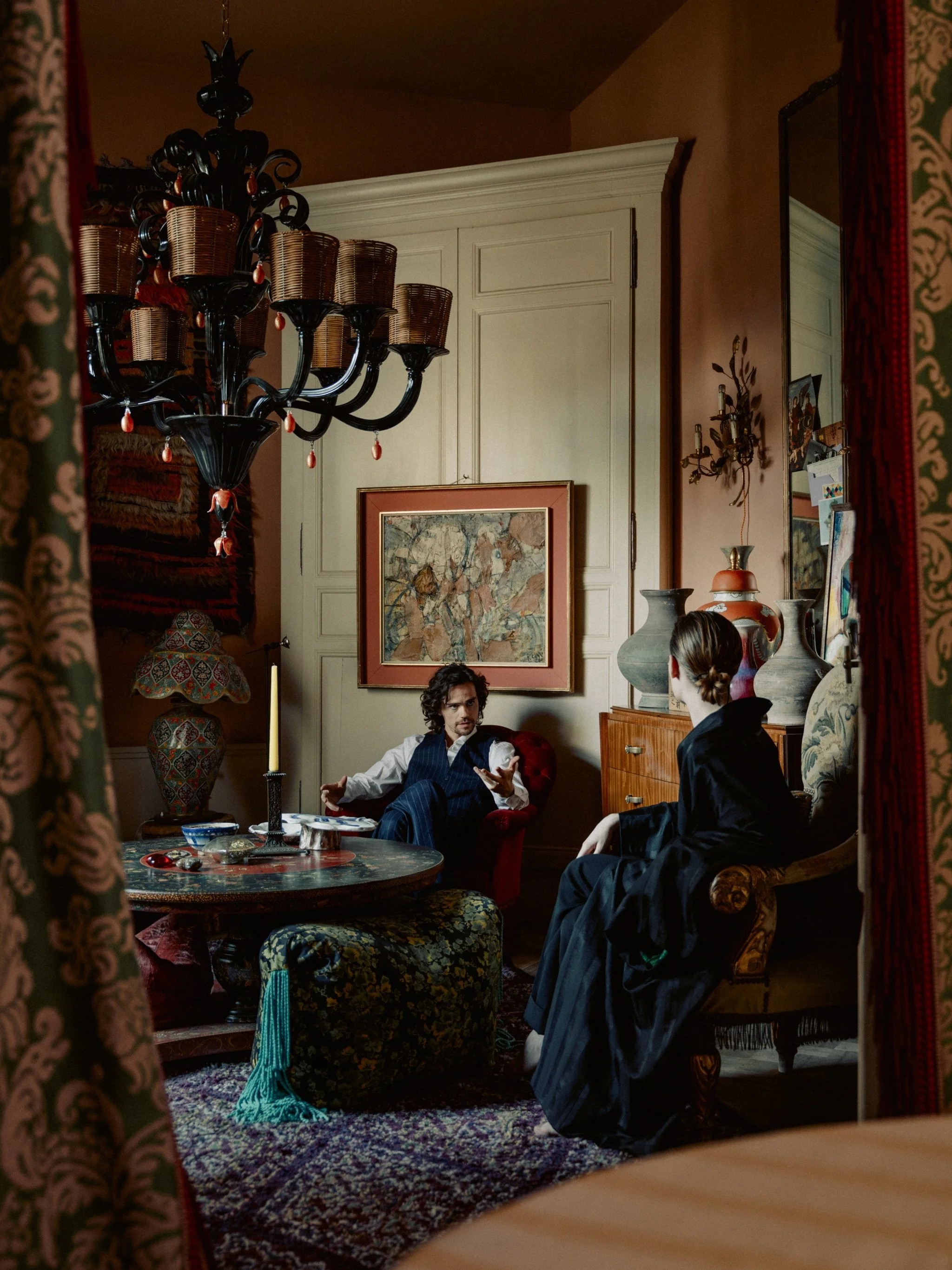 Two people sitting in a vintage decorated room, one with curly hair wearing a vest and the other with short hair in a long dress, engaged in conversation. The room features a chandelier, paintings, vases, a table with objects, and patterned furniture.