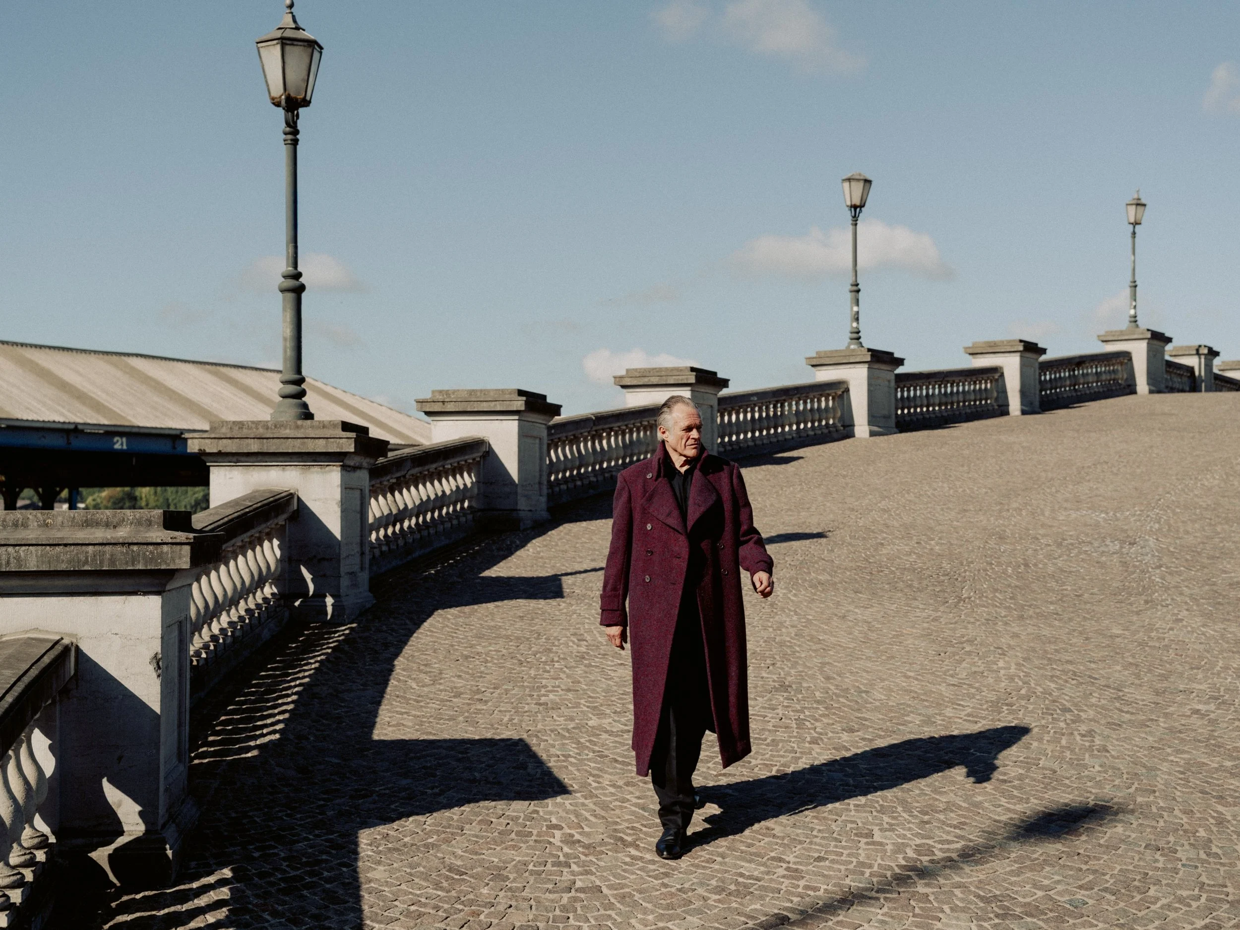 An elderly man with gray hair, wearing a long maroon coat, walks along a cobblestone path beside a decorative stone railing with street lamps, on a sunny day with a partly cloudy sky.