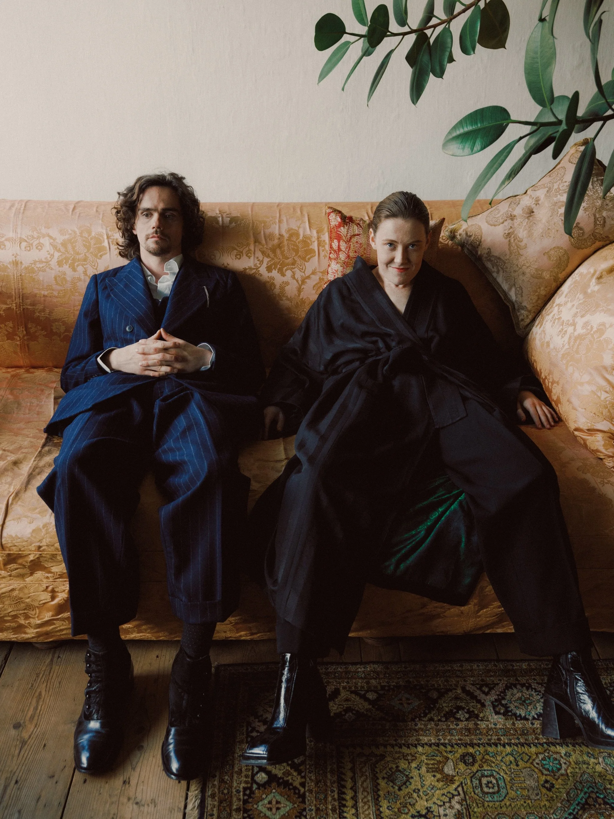 A man and woman sitting on a vintage floral-patterned sofa, dressed in stylish, dark, and patterned clothing.