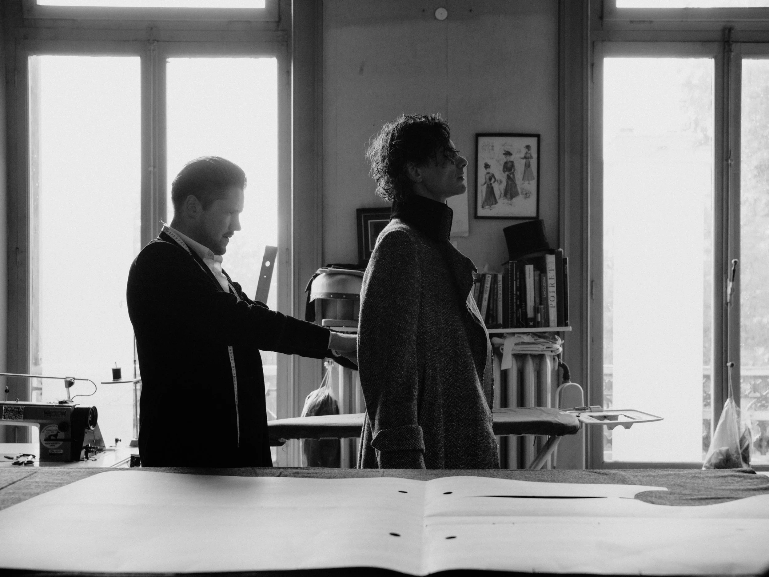 A person working on a sewing machine with another person standing nearby in a room with large windows and shelves of books.