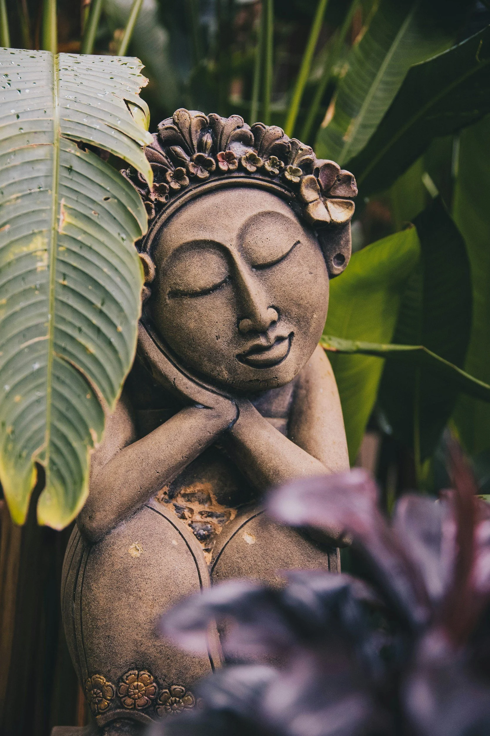 A stone statue of a woman with closed eyes, smiling, with her hands under her chin, surrounded by large green leaves.
