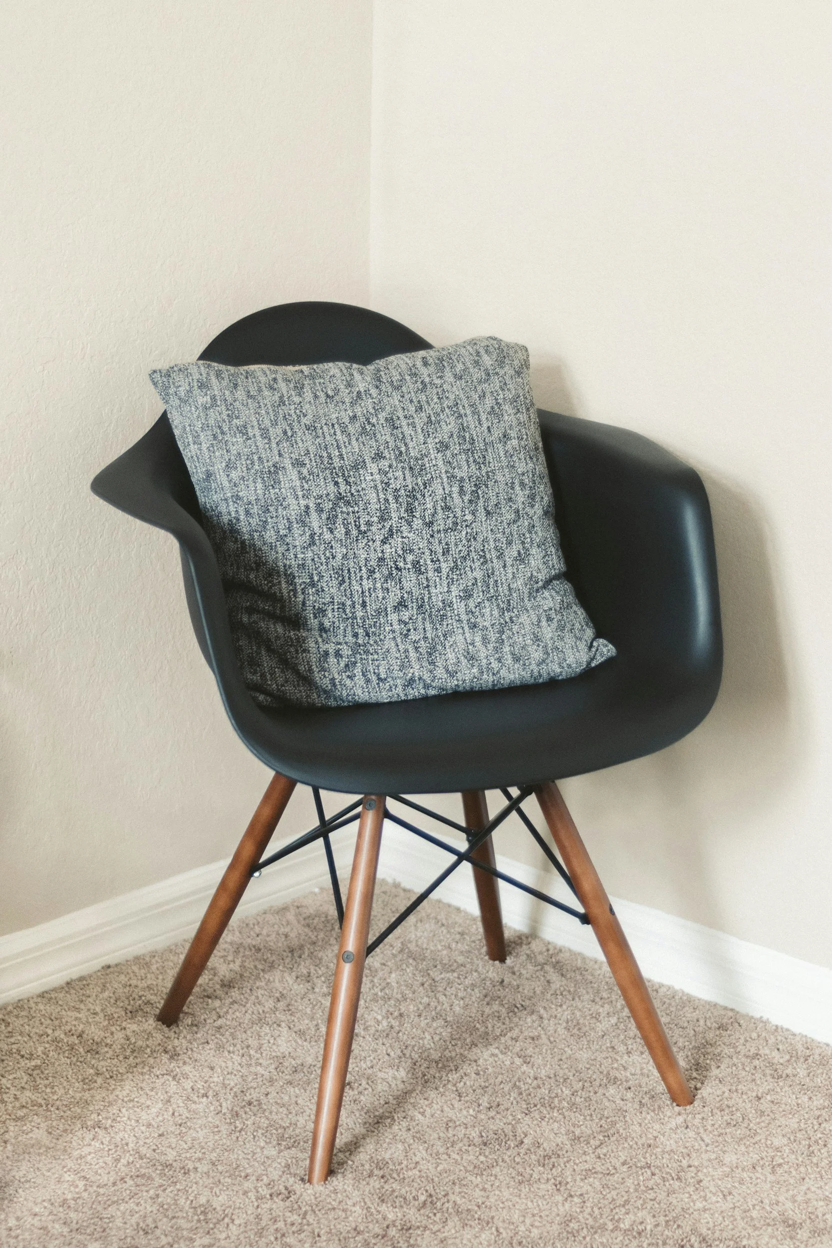 A black modern chair with wooden legs and a gray cushion placed on it, positioned in a corner next to beige walls and carpeted floor.