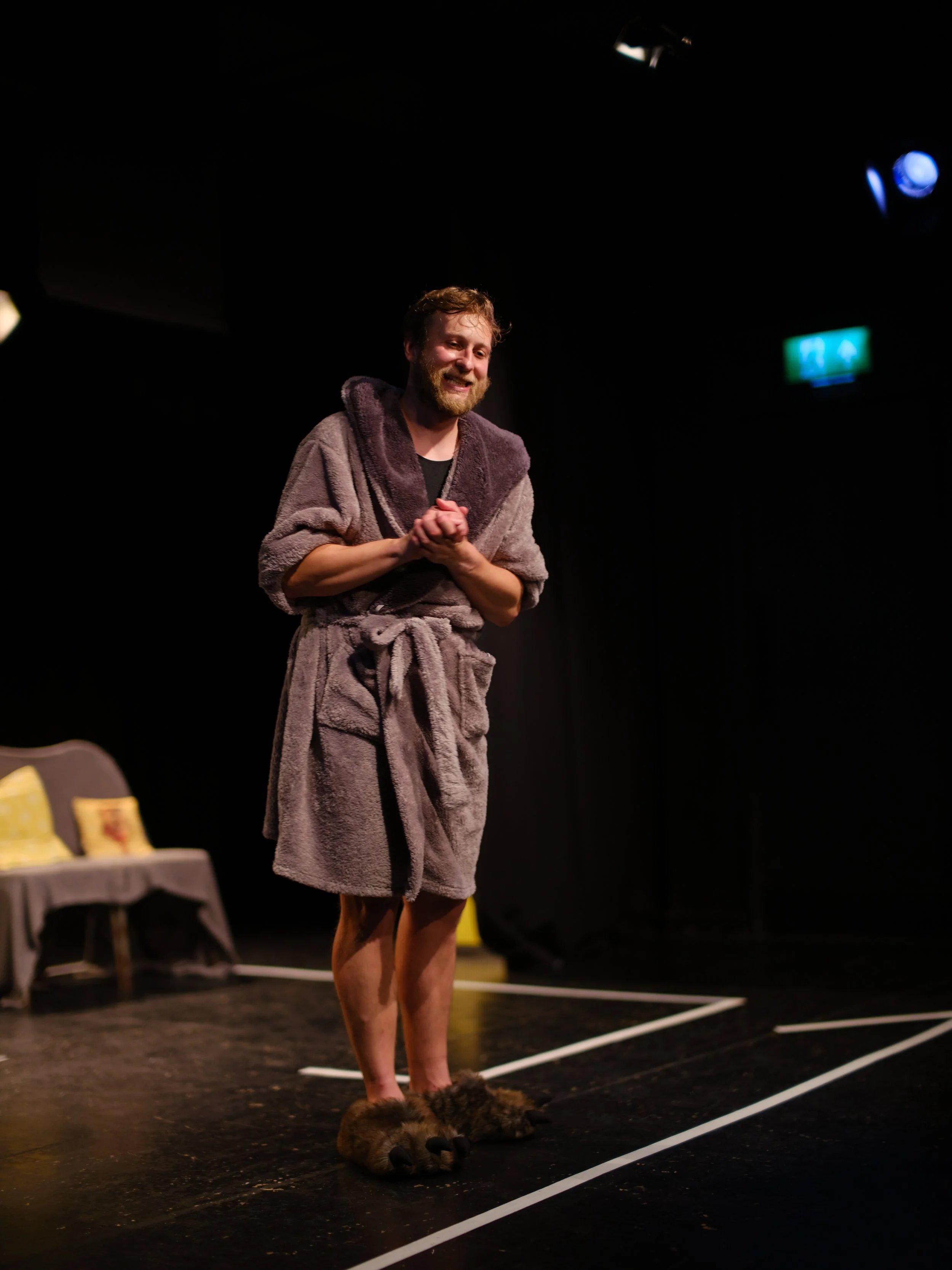 A man with a beard smiling on stage wearing a robe and animal slippers, standing in front of a dark background with stage lights.