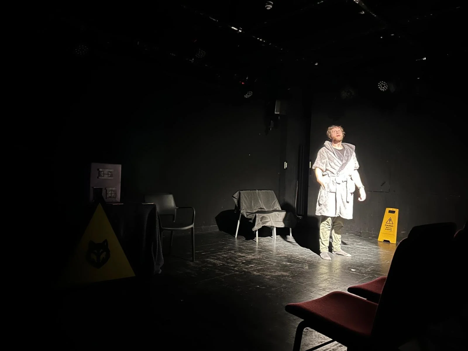 A person performing on a small stage illuminated by a spotlight, with black walls, a chair covered with a cloth, a table with a tablecloth, and a caution sign on the floor.